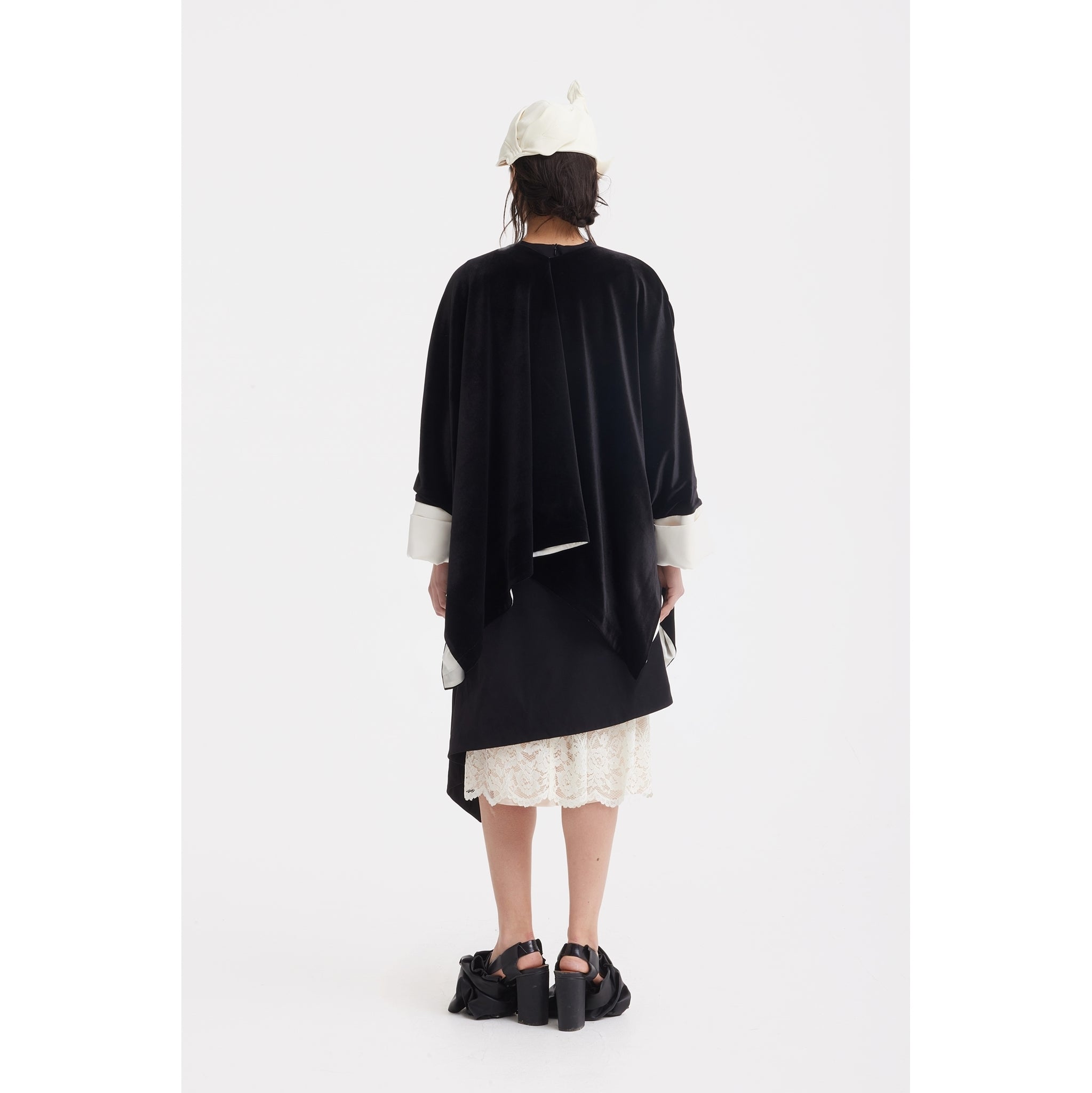 A person faces away, wearing the JULIA ALLERT Loose Velvet Cape Jacket With Zip Front in black with white cuffs, a lace skirt, white headscarf, and black platform sandals. The style highlights elegant layering against a plain white background.