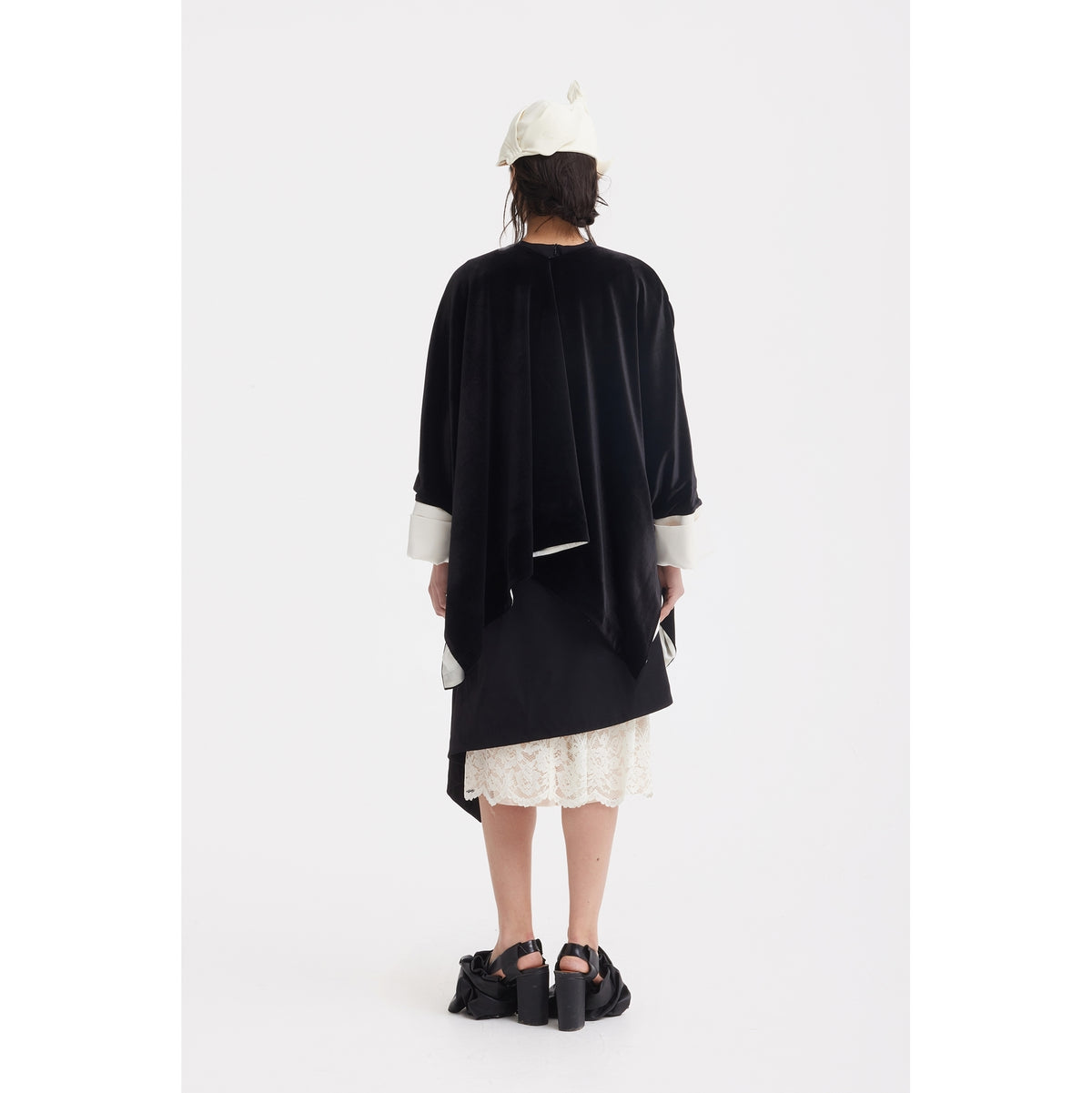 A person faces away, wearing the JULIA ALLERT Loose Velvet Cape Jacket With Zip Front in black with white cuffs, a lace skirt, white headscarf, and black platform sandals. The style highlights elegant layering against a plain white background.