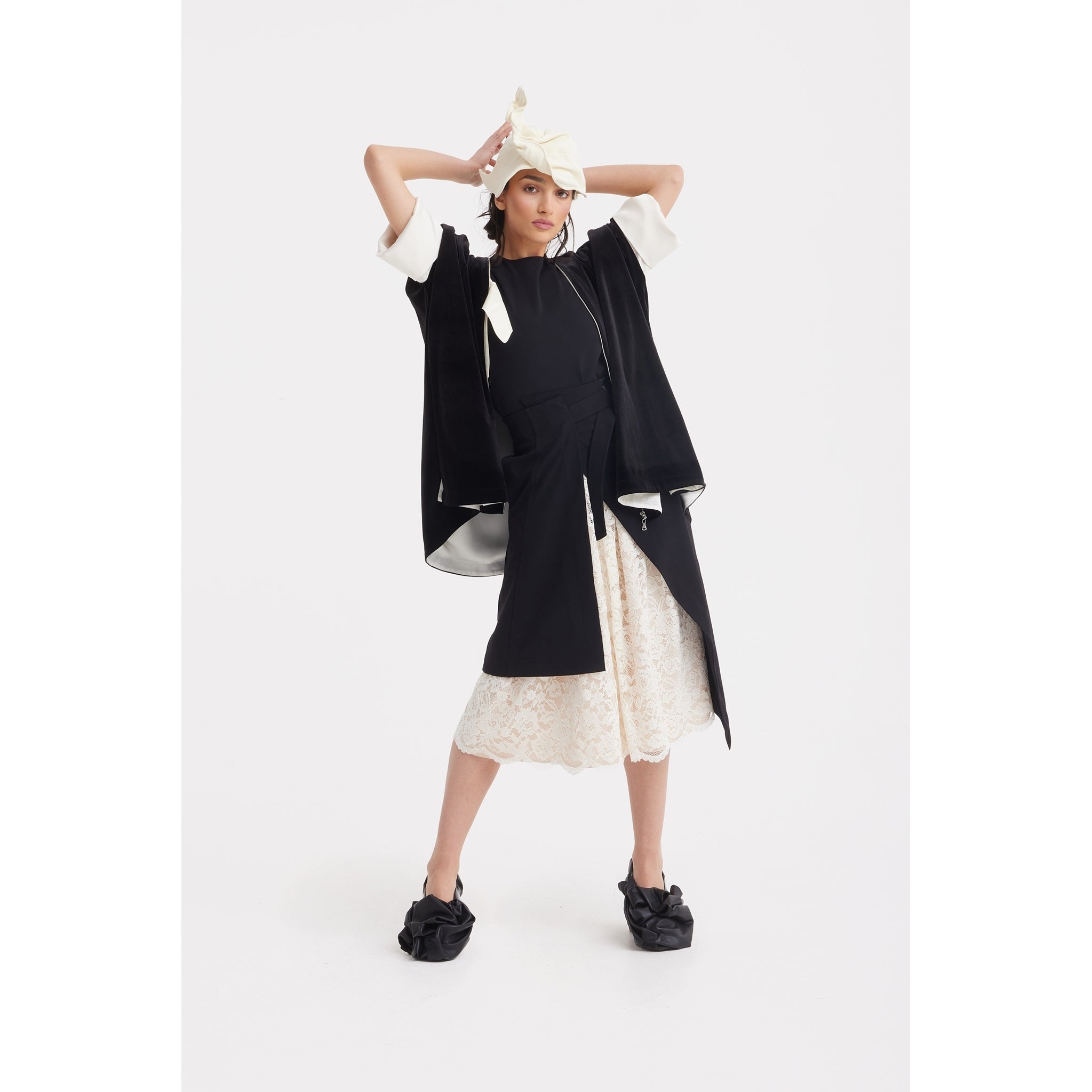 A person layers evening wear with JULIA ALLERT's Loose Velvet Cape Jacket With Zip Front in black, styled over a white lace skirt and black slip-on shoes. A cream headscarf completes the look against a plain white background.