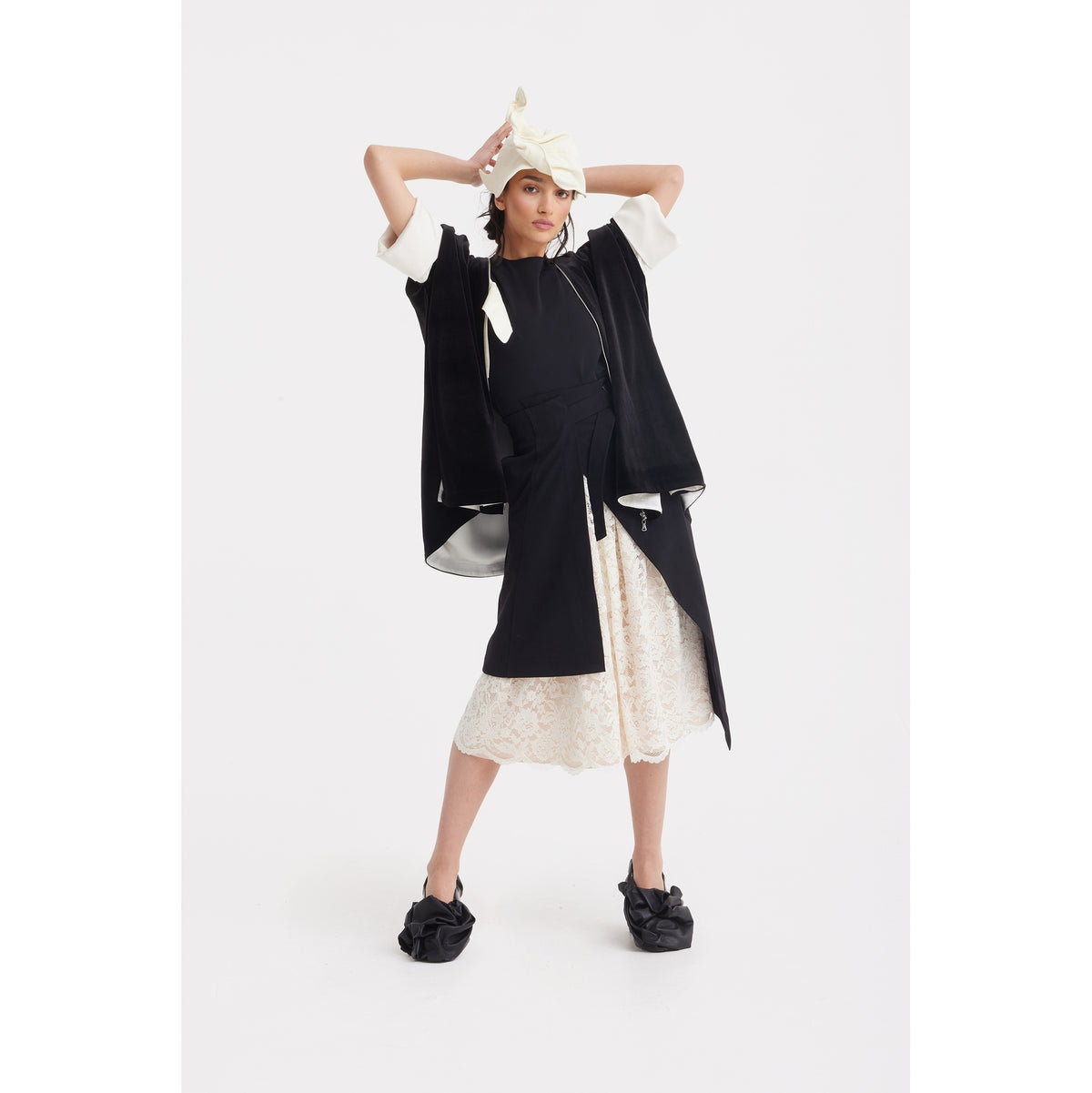 A person layers evening wear with JULIA ALLERT's Loose Velvet Cape Jacket With Zip Front in black, styled over a white lace skirt and black slip-on shoes. A cream headscarf completes the look against a plain white background.