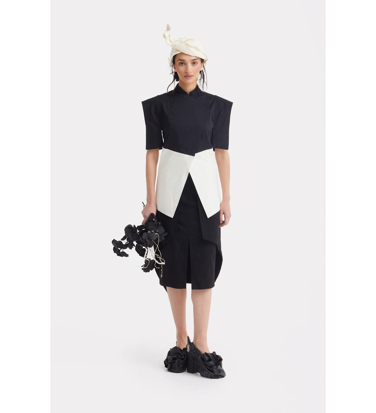 A woman stands against a plain white background wearing the JULIA ALLERT Asymmetric Colour Block Top in black &amp; white, accessorized with a large white belt or sash, cream beret, ruffled black shoes, and holding black flowers with ribbon details.