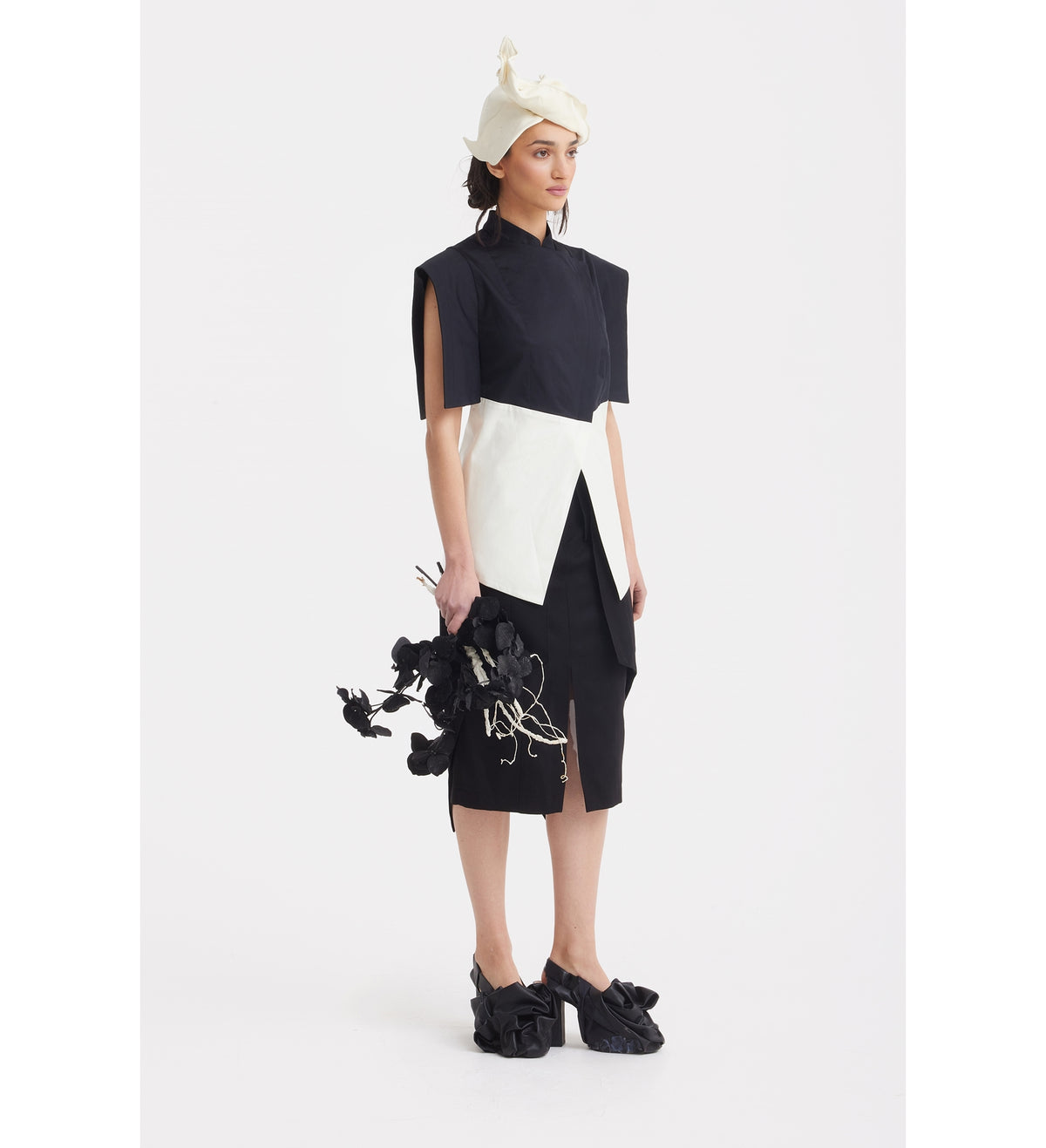 Against a white background, a person wears the JULIA ALLERT Asymmetric Colour Block Top in black &amp; white with a matching cream hat, holding black flowers and sporting sculptural black shoes for a striking modern look.