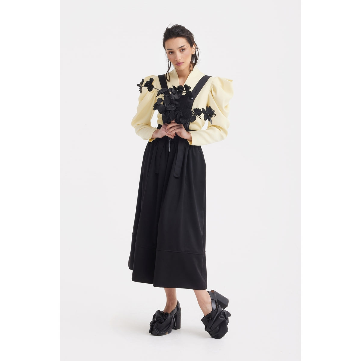 A woman poses against a white background wearing the JULIA ALLERT Sculpted V-Neck Blouse with Dramatic Sleeves in pale yellow, paired with a long black skirt and black bow-adorned shoes, hands together at her waist.