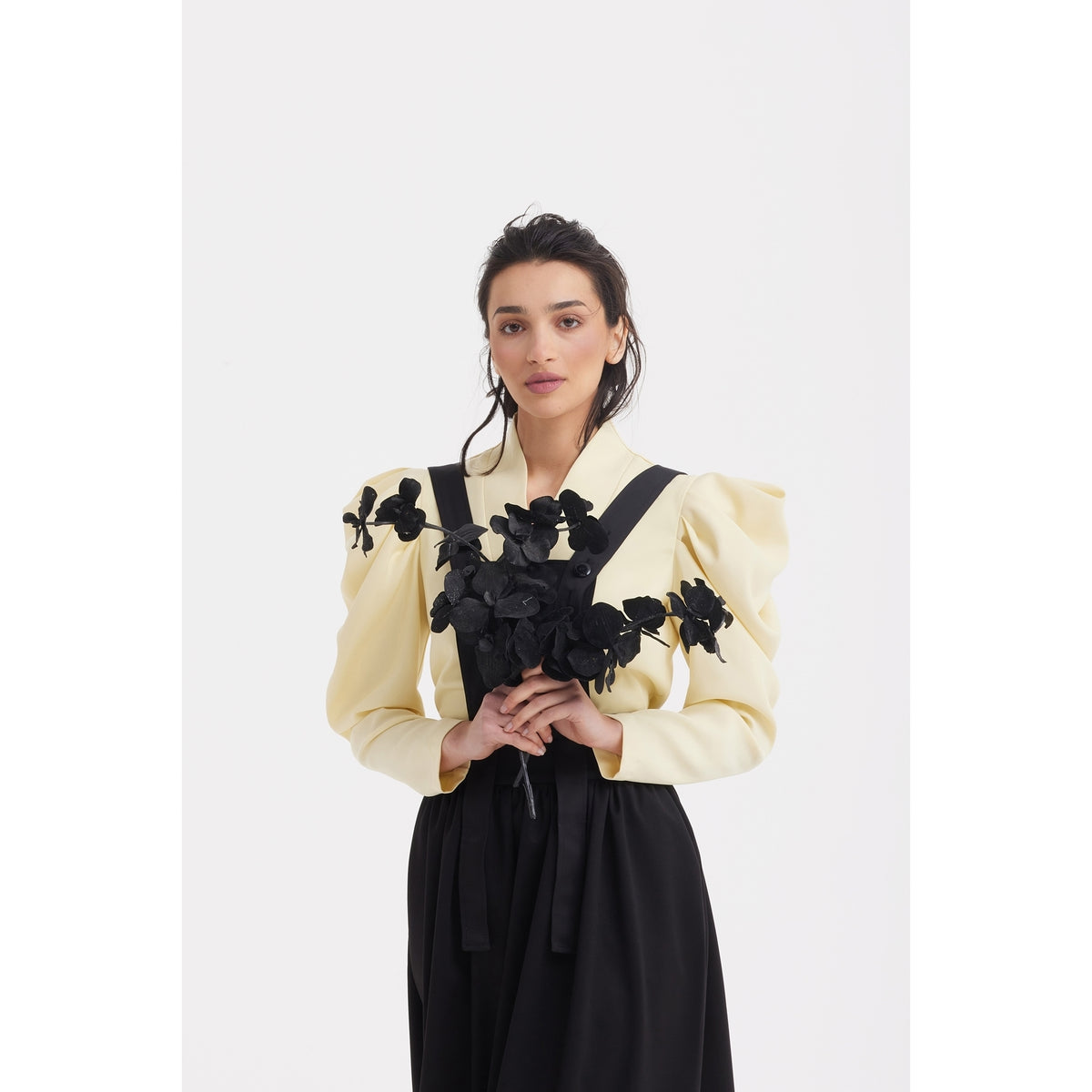A woman wears the JULIA ALLERT Sculpted V-Neck Blouse with Dramatic Sleeves in pale yellow, adorned with black floral details. She holds a bouquet of black flowers and pairs the blouse with a structured black suiting skirt.