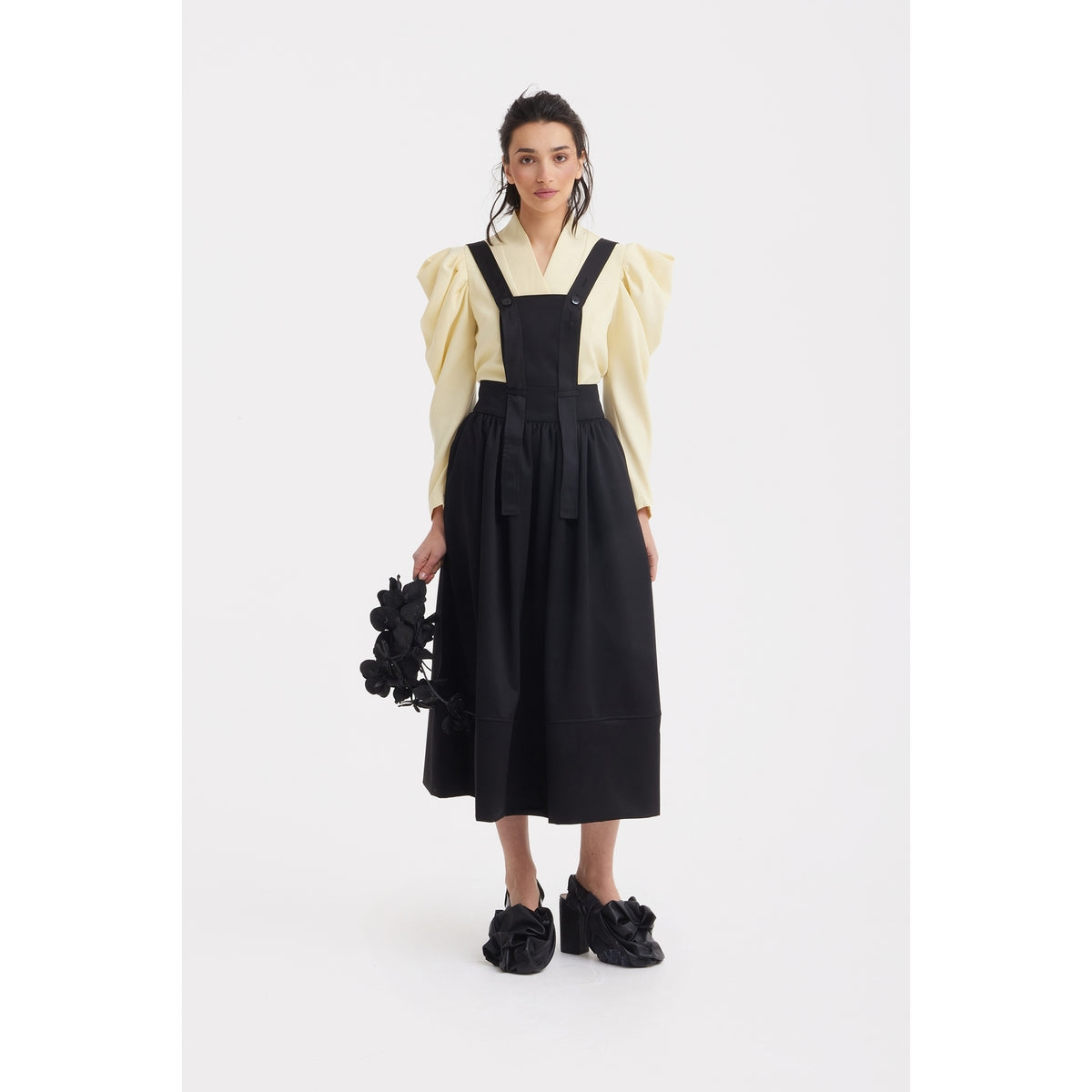 A woman stands facing forward in a JULIA ALLERT Sculpted V-Neck Blouse with Dramatic Sleeves in pale yellow under a black pinafore, holding a black floral accessory and wearing black shoes with large fabric bows against a white background.