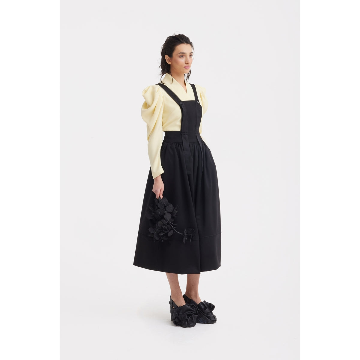 A woman stands against a white background wearing the JULIA ALLERT Sculpted V-Neck Blouse with Dramatic Sleeves in pale yellow under a black pinafore dress, holding a black floral accessory and wearing black shoes with large decorative flowers.