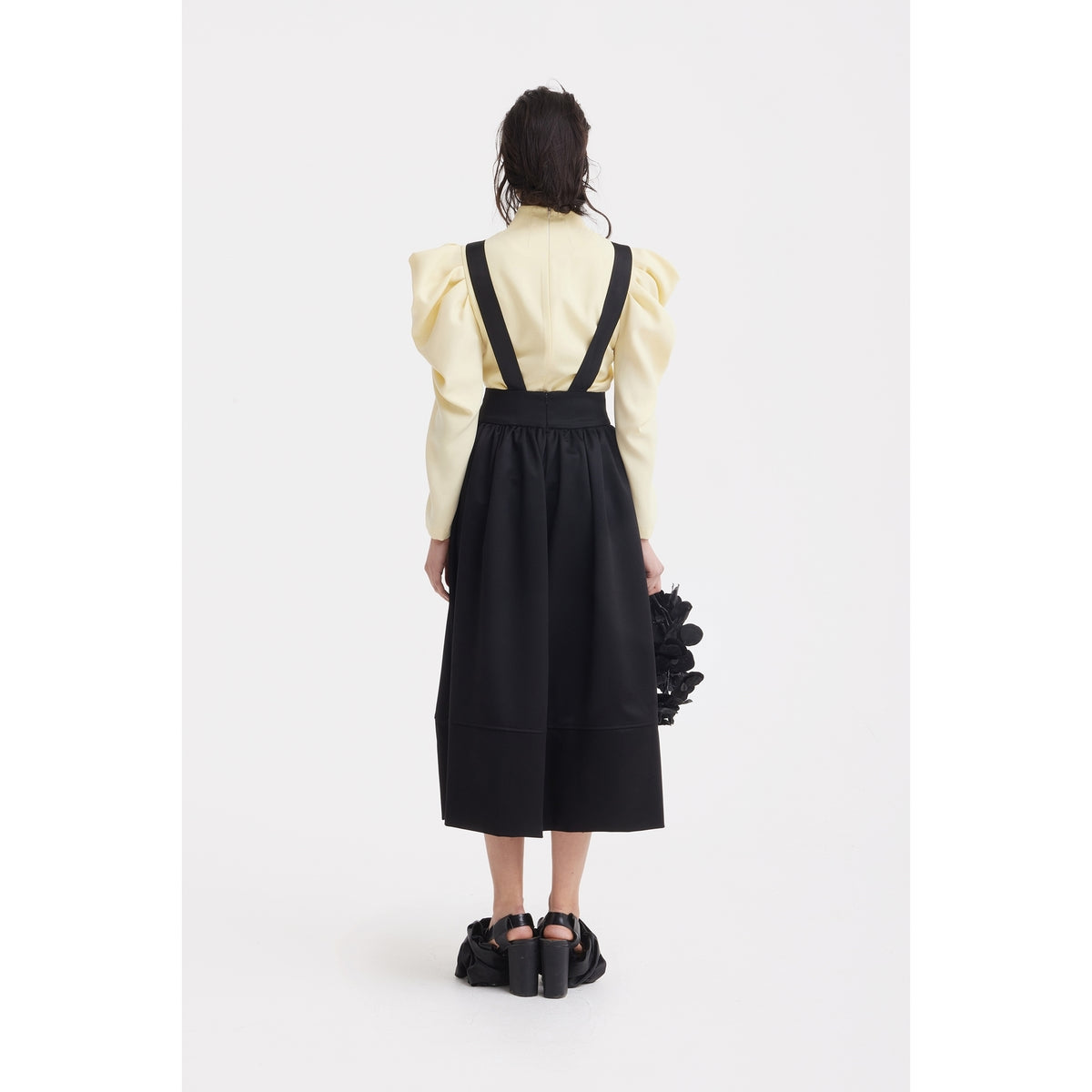 A person faces away, wearing the JULIA ALLERT Sculpted V-Neck Blouse with Dramatic Sleeves in pale yellow and a black suspender skirt. They hold a black textured bag and wear black shoes against a plain white background.
