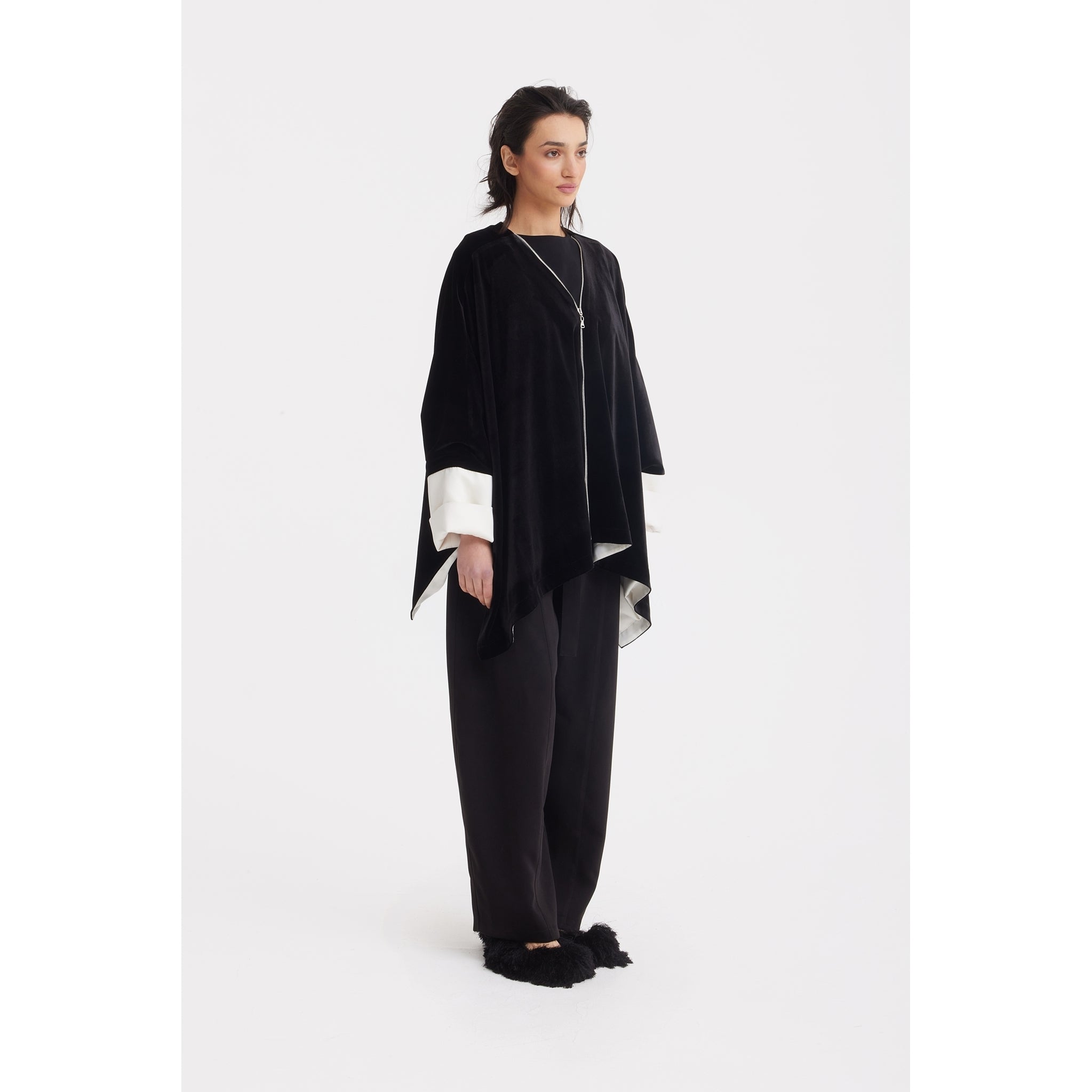 A woman stands against a white background wearing the JULIA ALLERT Loose Velvet Cape Jacket With Zip Front in black, paired with loose black pants and black furry slippers—her dark hair pulled back as she gazes slightly to the side.