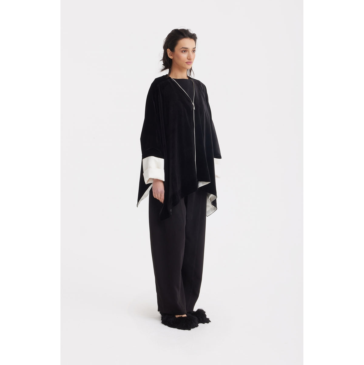 A woman stands against a white background wearing the JULIA ALLERT Loose Velvet Cape Jacket With Zip Front in black, paired with loose black pants and black furry slippers—her dark hair pulled back as she gazes slightly to the side.