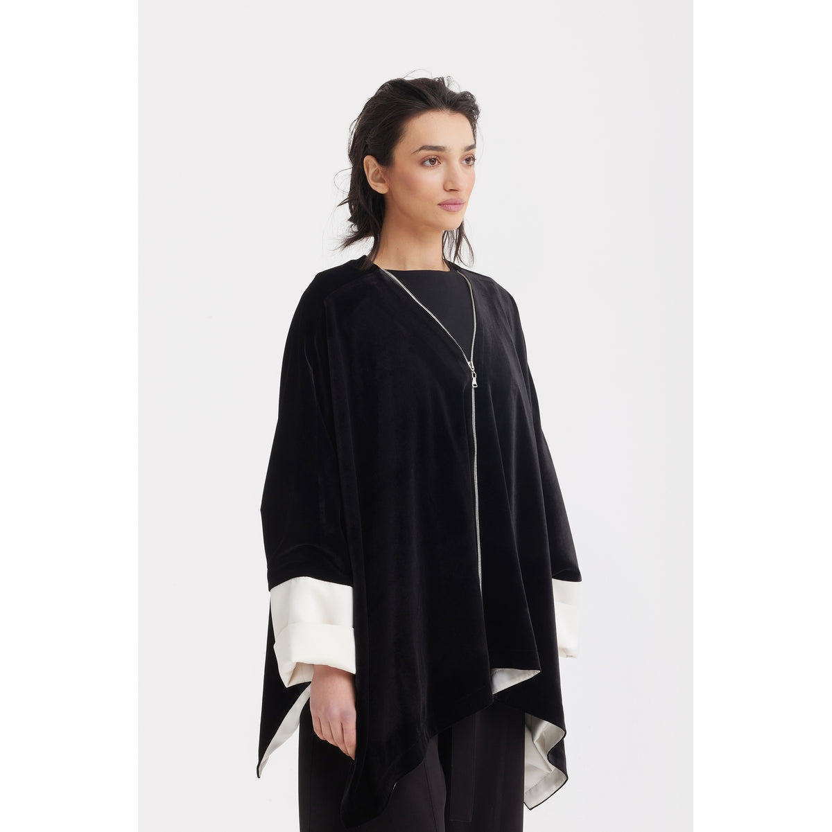A woman models the JULIA ALLERT Loose Velvet Cape Jacket With Zip Front in black, layered over a black top. She stands against a plain white background, looking to the side with a neutral expression.