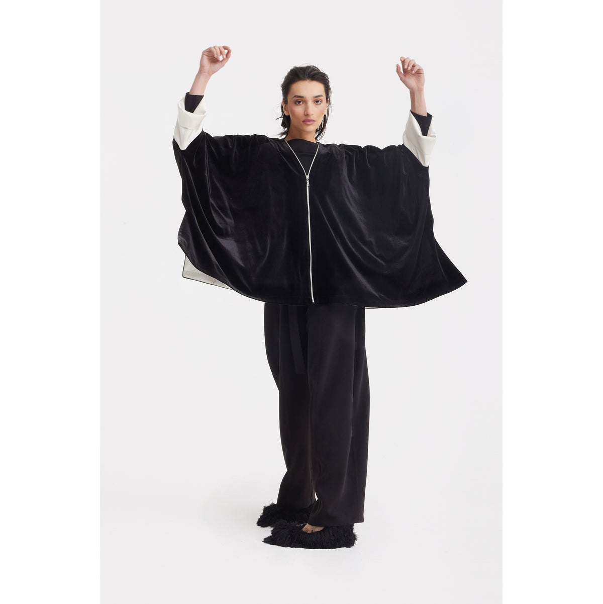 A person stands with arms raised, modeling the JULIA ALLERT Loose Velvet Cape Jacket With Zip Front in black—an elegant evening layering piece with white sleeves and loose black pants, set against a plain white background.