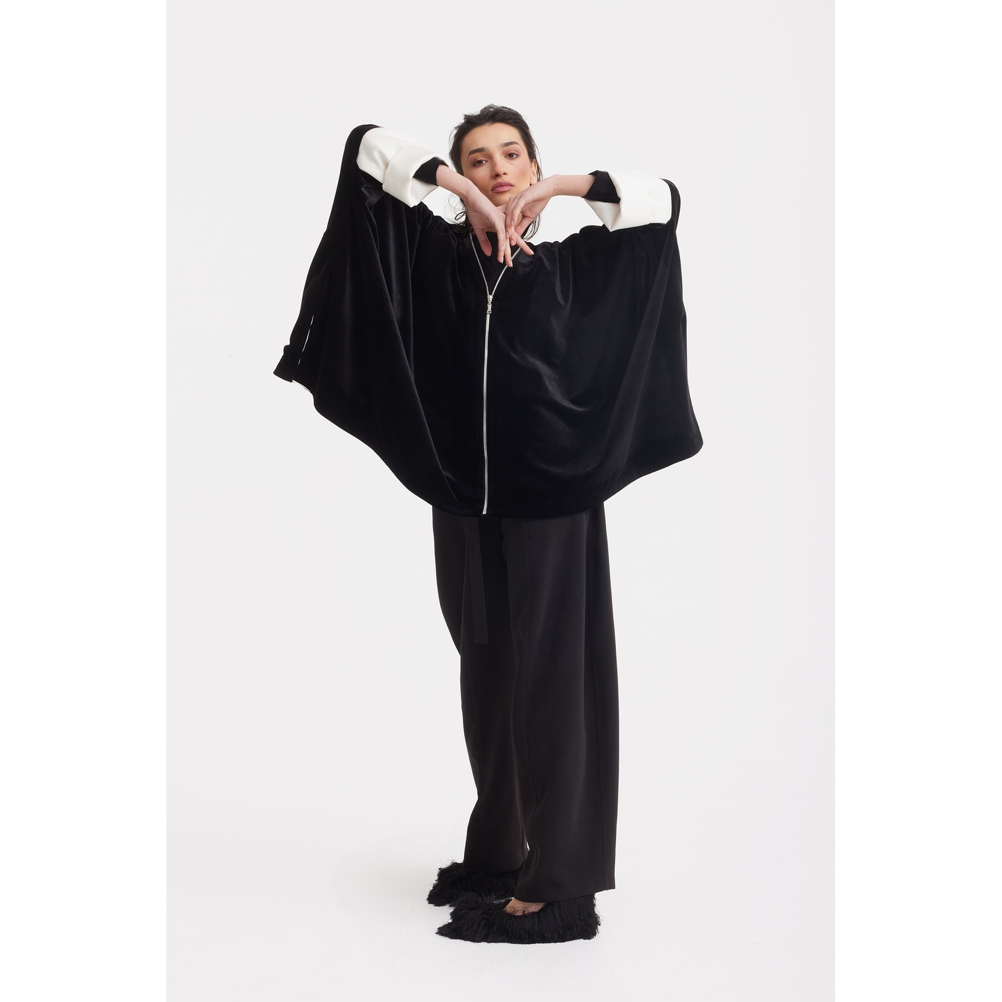 A woman poses against a white background in the JULIA ALLERT Loose Velvet Cape Jacket With Zip Front - Black, featuring wide white cuffs, styled with wide-leg black trousers and black shoes. She raises her arms, hands together at chin—ideal for elegant evenings.