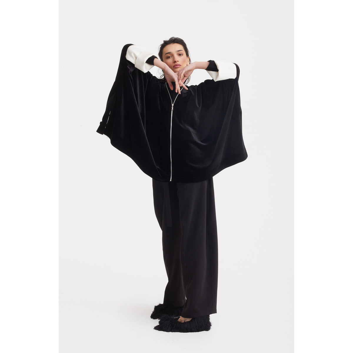 A woman poses against a white background in the JULIA ALLERT Loose Velvet Cape Jacket With Zip Front - Black, featuring wide white cuffs, styled with wide-leg black trousers and black shoes. She raises her arms, hands together at chin—ideal for elegant evenings.