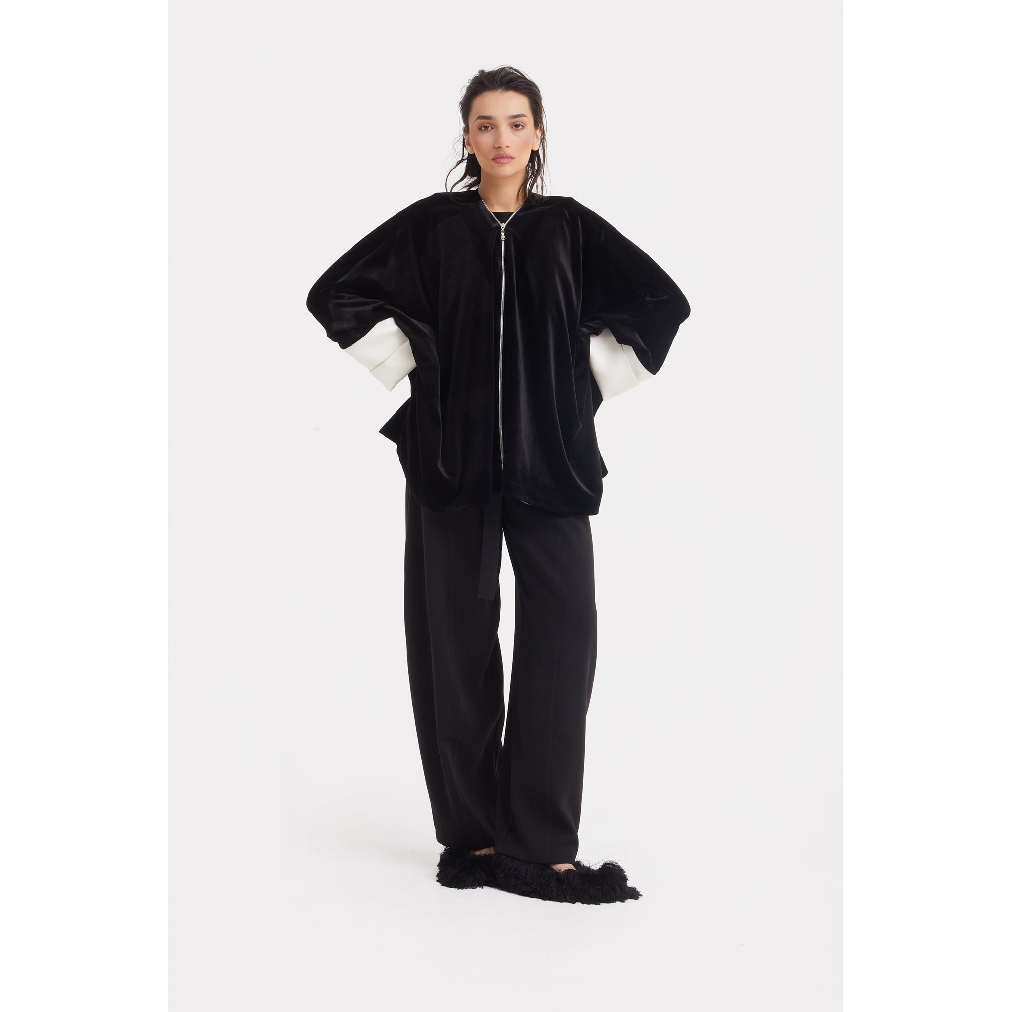 A woman poses against a white background in the JULIA ALLERT Loose Velvet Cape Jacket With Zip Front - Black, styled over a white shirt with black wide-leg pants and furry black slippers—ideal for chic, layered evening looks.