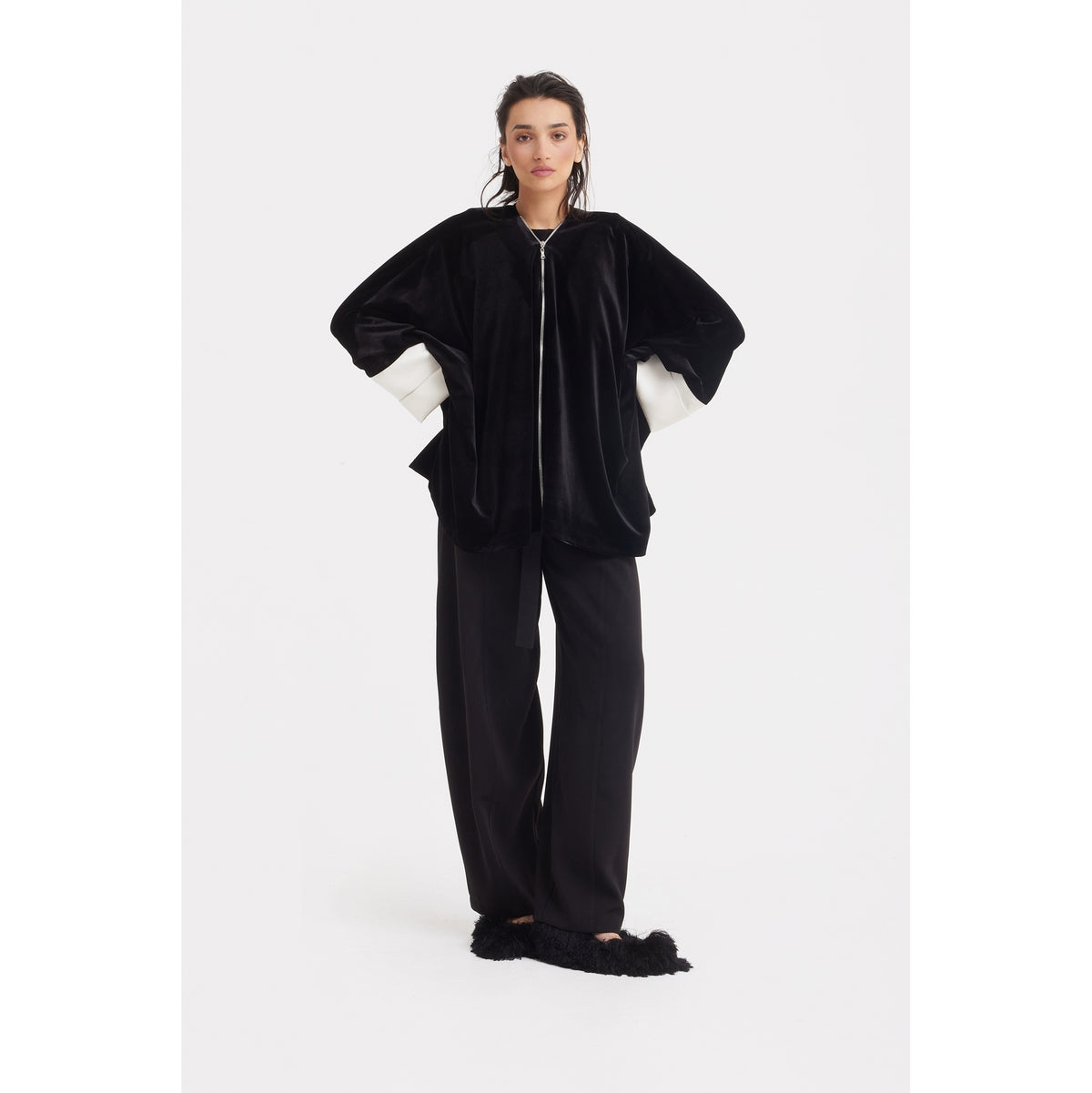 A woman poses against a white background in the JULIA ALLERT Loose Velvet Cape Jacket With Zip Front - Black, styled over a white shirt with black wide-leg pants and furry black slippers—ideal for chic, layered evening looks.
