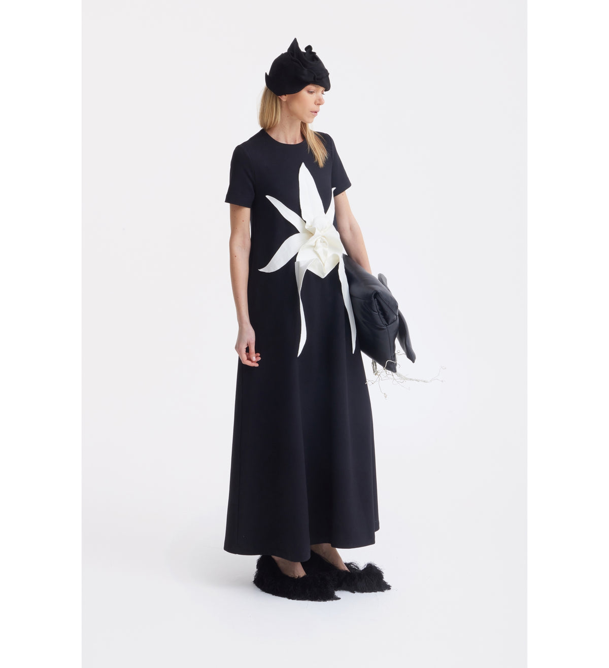 A woman poses against a white background in the Ankle-Length Knit Dress With Flower Detail - Black by JULIA ALLERT. The dress features short sleeves and a sculptural floral appliqué. She accessorizes with black fuzzy shoes, a black headpiece, and holds a jacket.