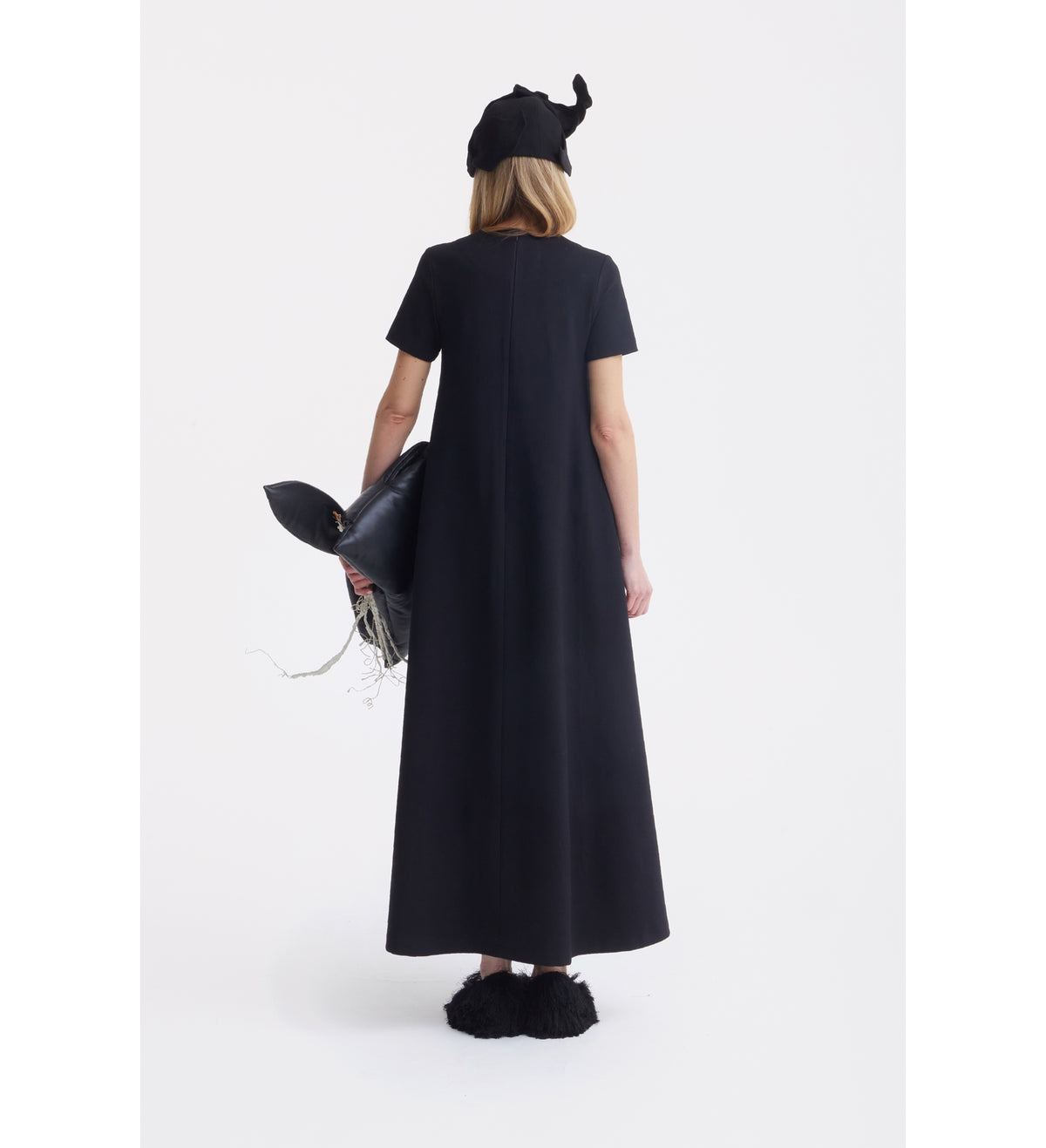 A woman faces away, wearing a JULIA ALLERT Ankle-Length Knit Dress With Flower Detail in black, a black hat, and black fuzzy shoes. She holds a large whale-shaped black bag with white string details against a plain white background.