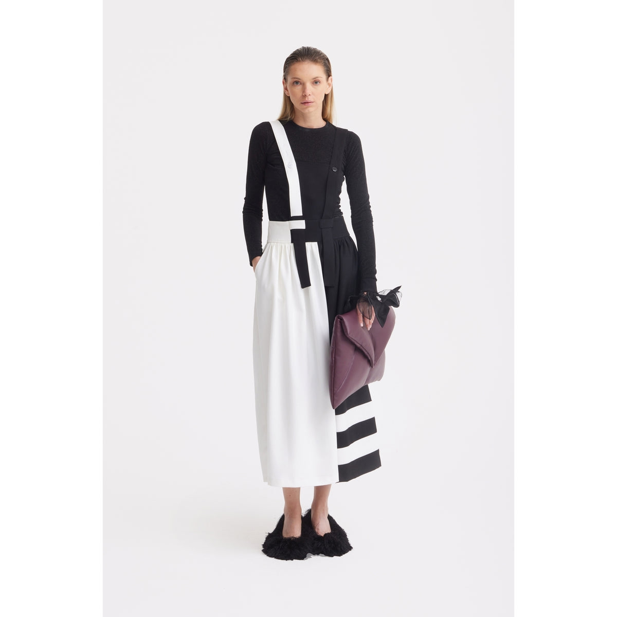 A woman stands against a white background wearing the JULIA ALLERT Midi Apron Dress With Adjustable Straps - Black Ivory, paired with a black long-sleeve top, black fuzzy shoes, and holding a large purple bag. Her straight hair is pulled back.