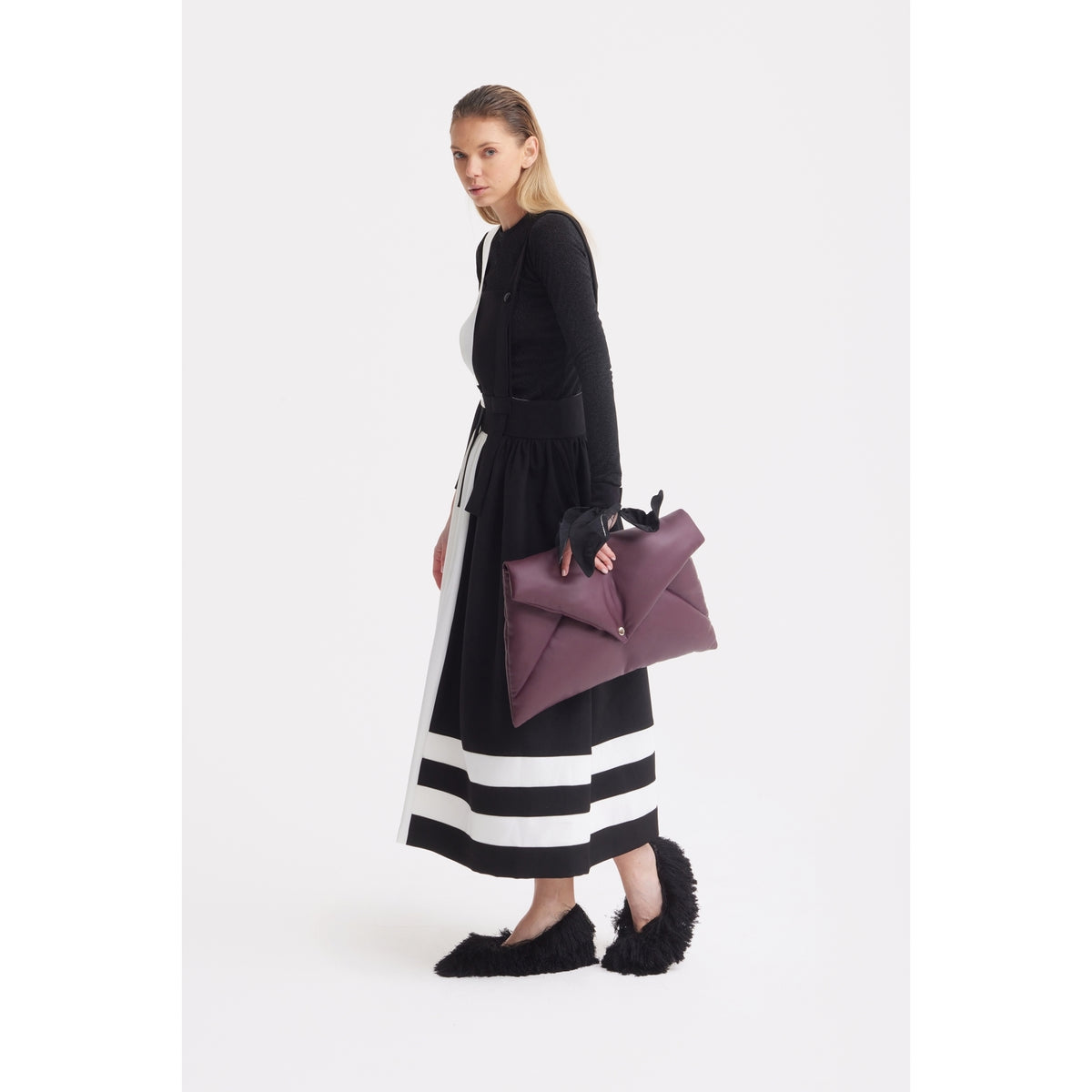 A woman wears the JULIA ALLERT Midi Apron Dress With Adjustable Straps in Black Ivory, featuring white striped hems, paired with a black long-sleeve top and fuzzy slippers, holding a large maroon handbag against a plain white background.