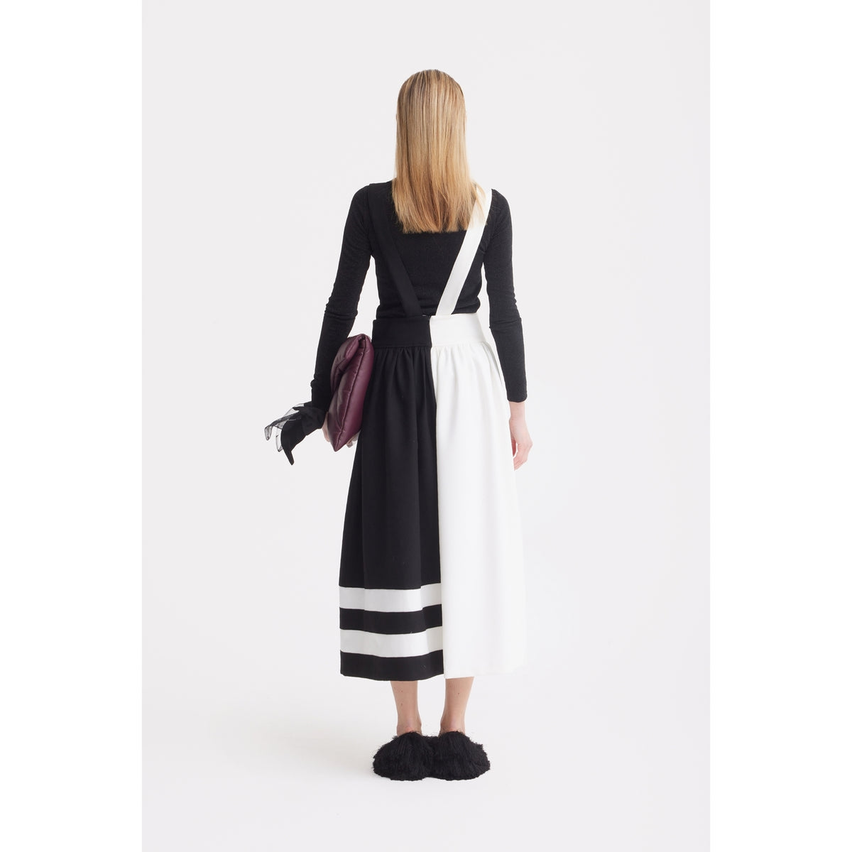 A woman with straight blonde hair, facing away against a white background, wears a black long-sleeve top and the JULIA ALLERT Midi Apron Dress With Adjustable Straps - Black Ivory, with a bag and black fluffy slippers.