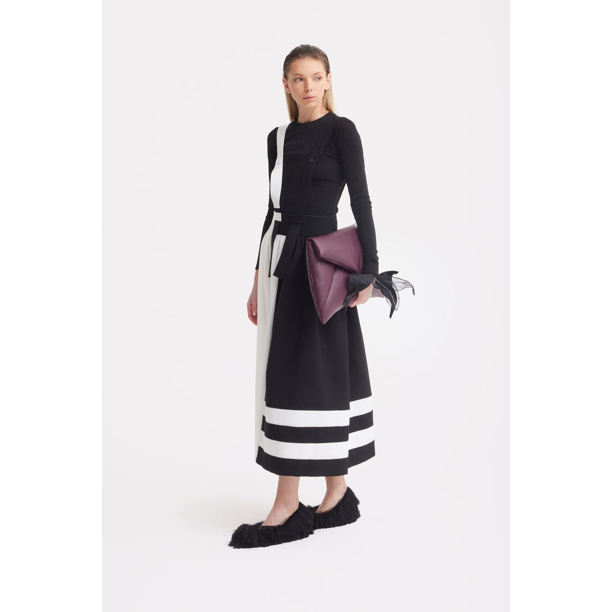 A woman with straight hair and a neutral expression stands against a white background wearing the JULIA ALLERT Midi Apron Dress With Adjustable Straps - Black Ivory, black textured shoes, and holding a large maroon clutch with black detailing.