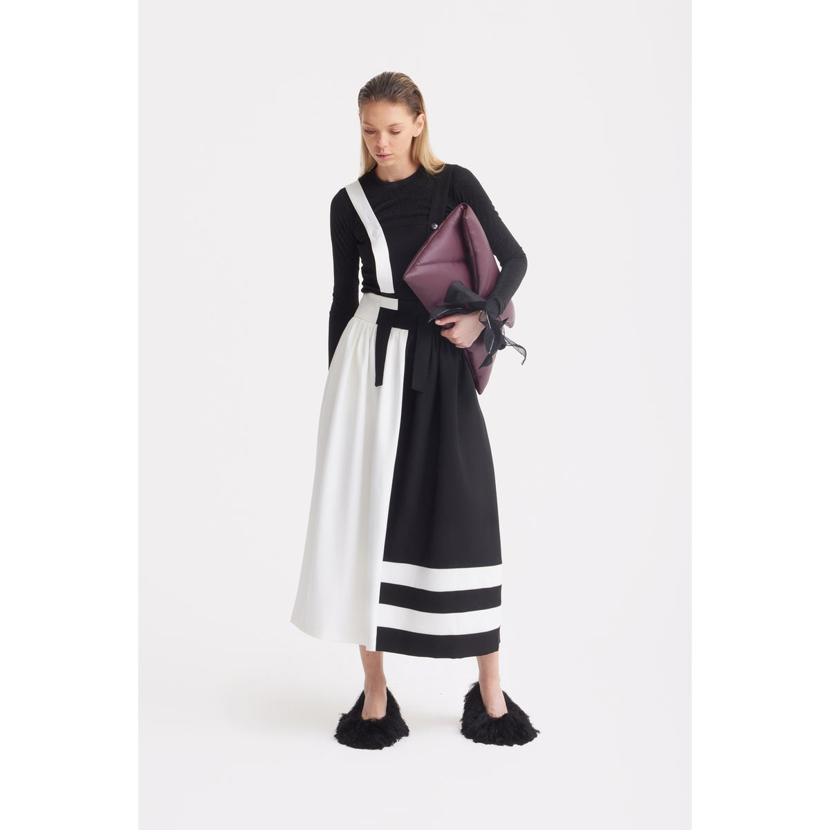 A woman stands against a plain white background wearing the JULIA ALLERT Midi Apron Dress With Adjustable Straps in Black Ivory, paired with a black long-sleeve top, black furry shoes, and holding a large purple clutch under her arm.