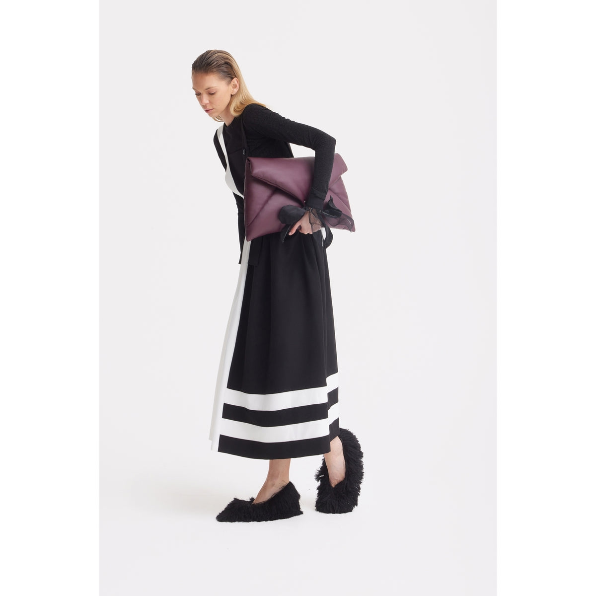 A woman wears the JULIA ALLERT Midi Apron Dress With Adjustable Straps in Black Ivory, paired with a black top and fuzzy slippers, holding a maroon clutch with a black ribbon against a plain white background.