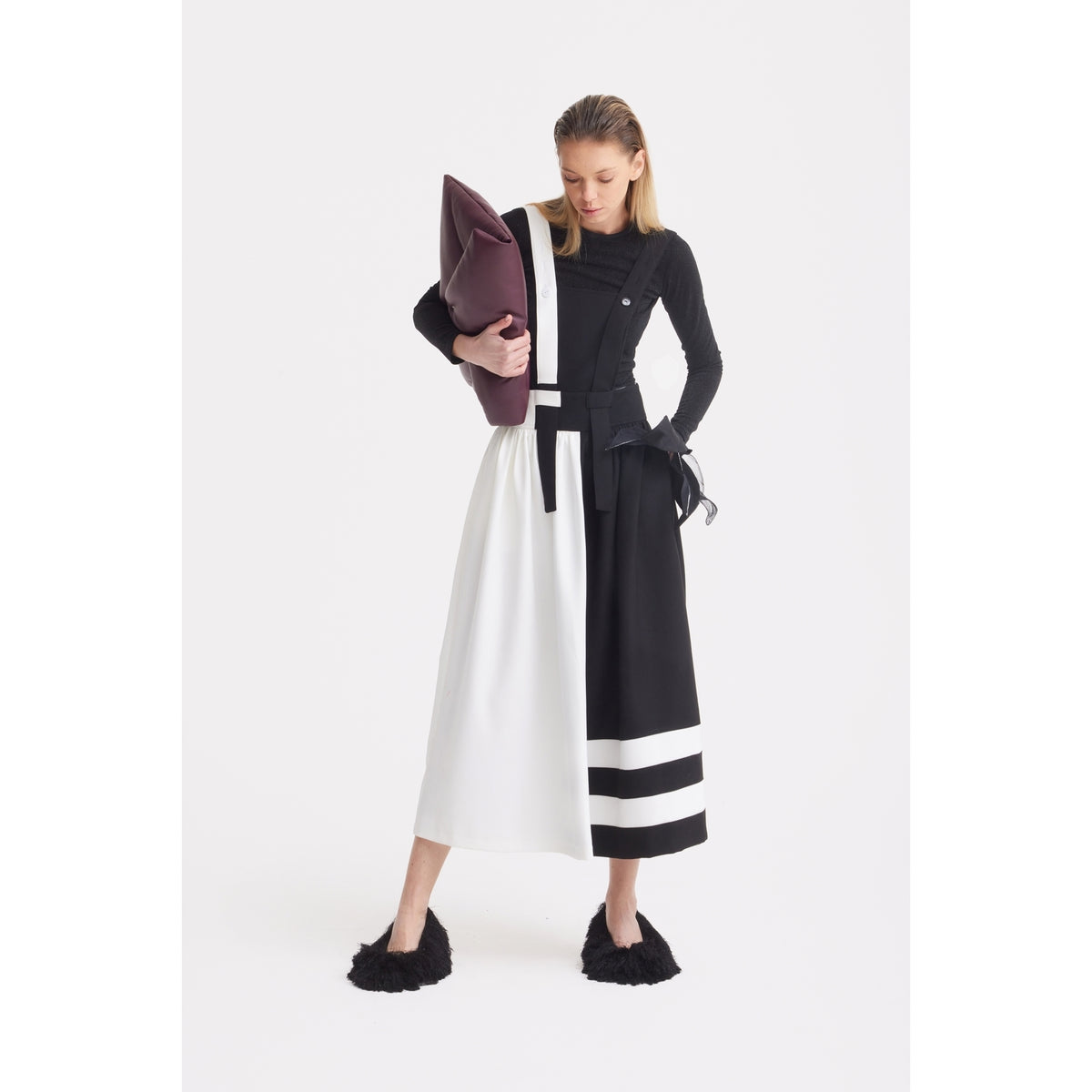 A woman stands against a white background wearing the JULIA ALLERT Midi Apron Dress With Adjustable Straps in Black Ivory, paired with a fitted black top, black furry slippers, and holding a large folded burgundy clutch under her arm.