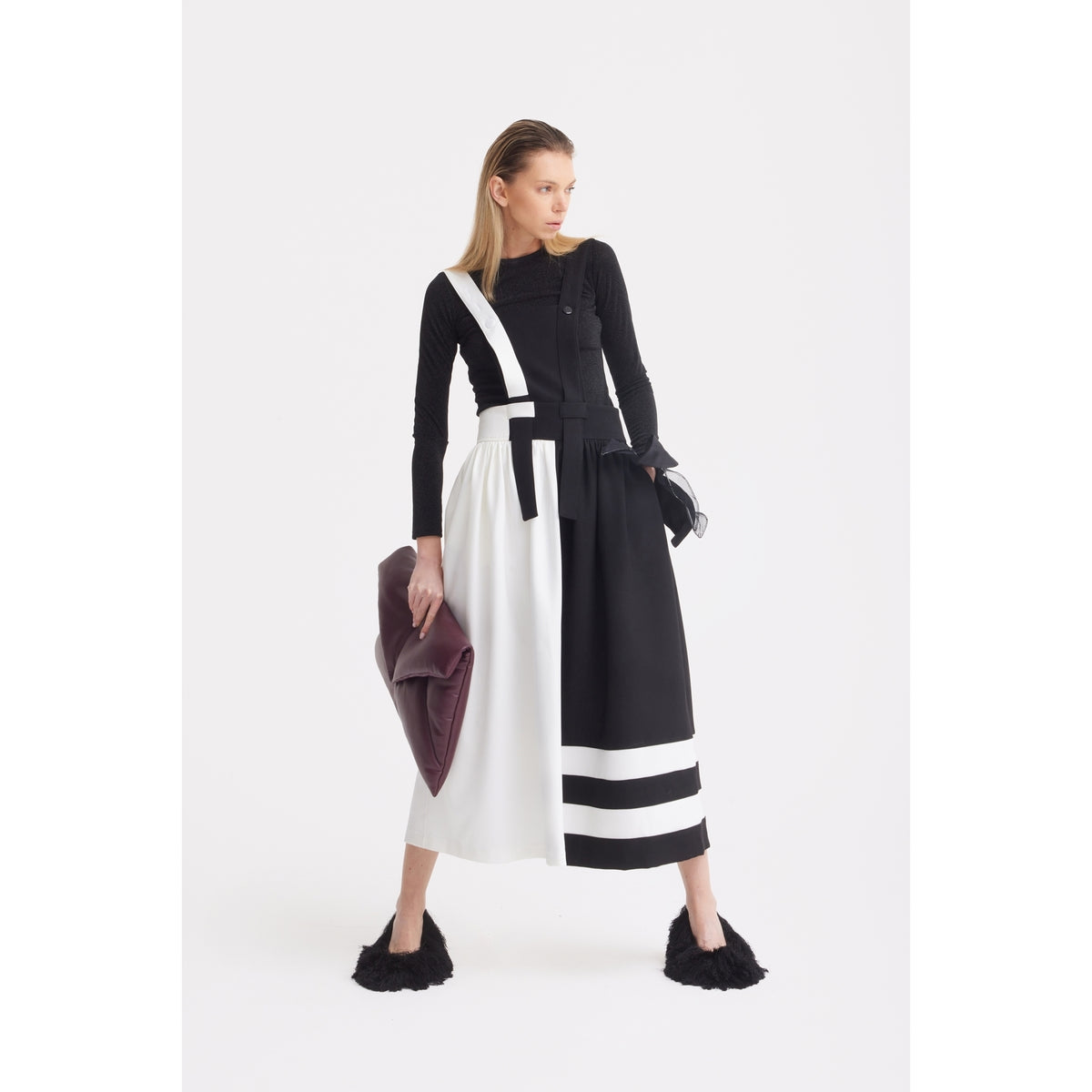 A woman stands against a white background wearing the JULIA ALLERT Midi Apron Dress With Adjustable Straps in Black Ivory, paired with black fuzzy shoes and holding a large burgundy clutch, gazing to the side confidently.