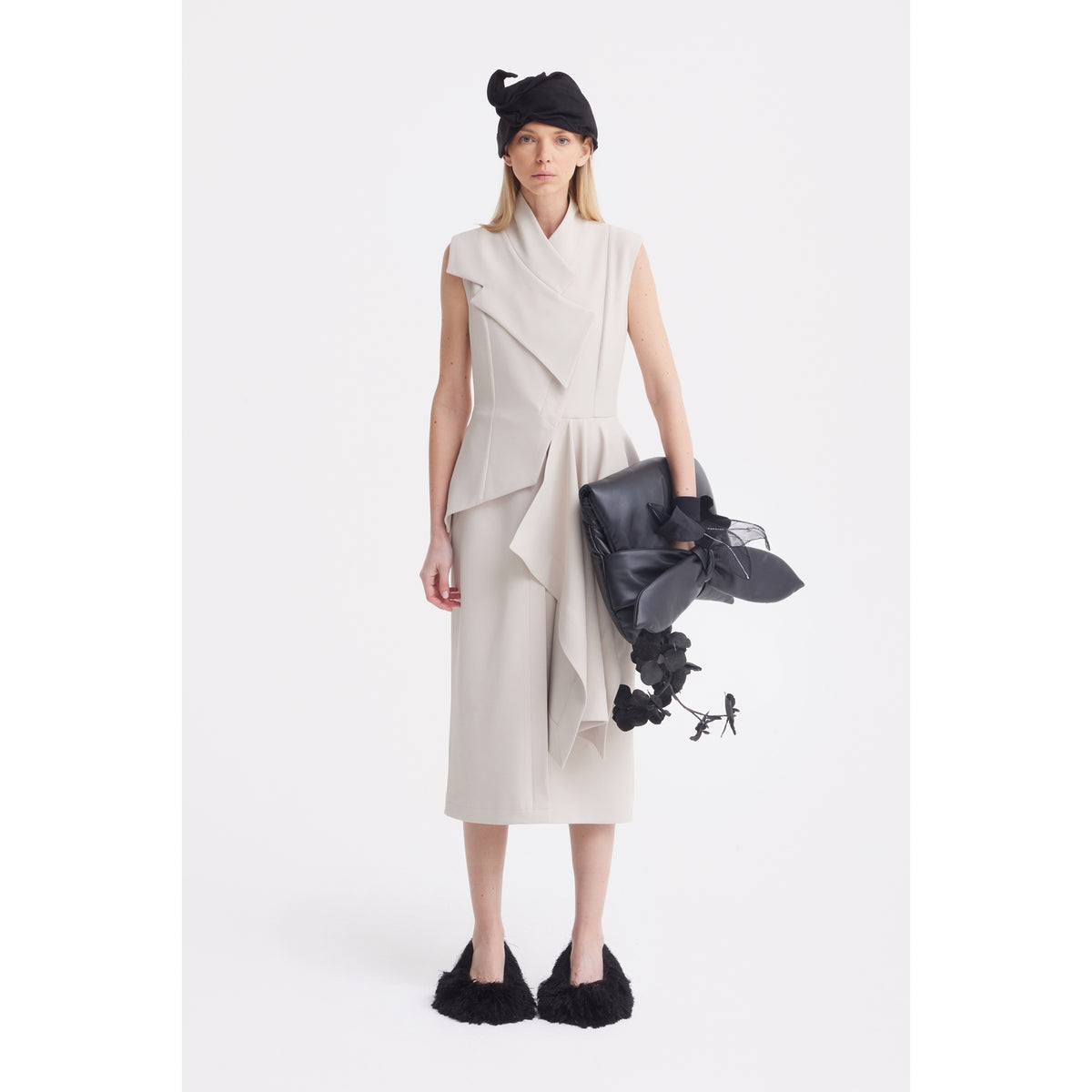 A woman faces forward in a JULIA ALLERT Architectural Draped Midi Dress in beige, paired with black furry shoes, a black beret-style hat, and holding a large sculptural black bag with floral accents against a plain white background.