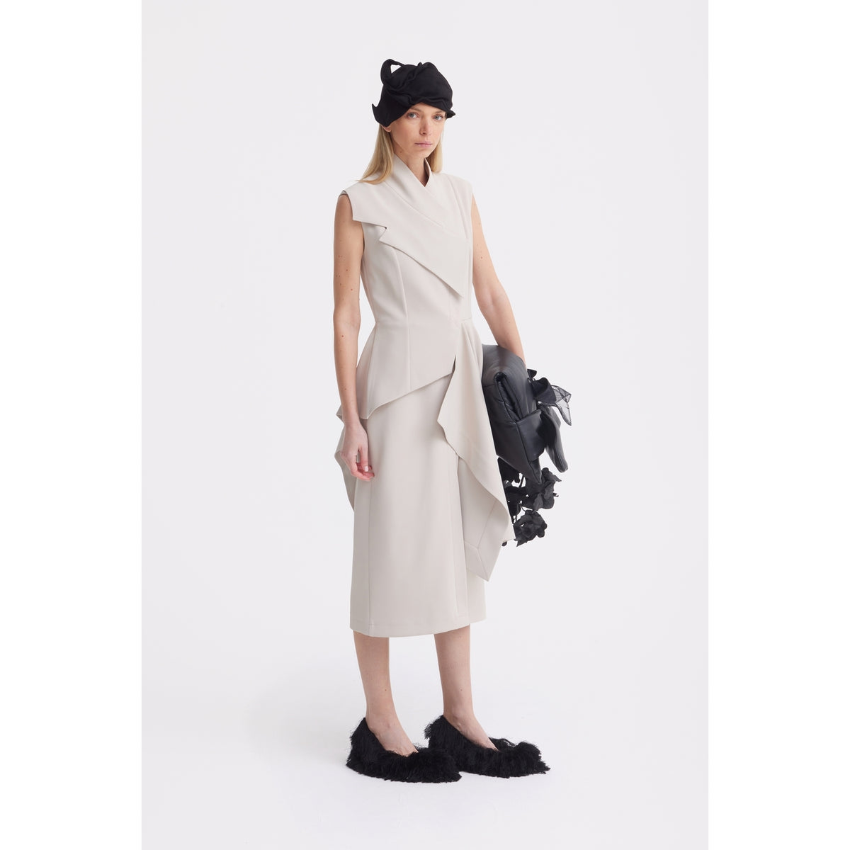 A woman stands against a plain white background wearing the JULIA ALLERT Architectural Draped Midi Dress in beige, paired with a black headpiece, black furry slip-on shoes, and holding a sculptural black accessory.
