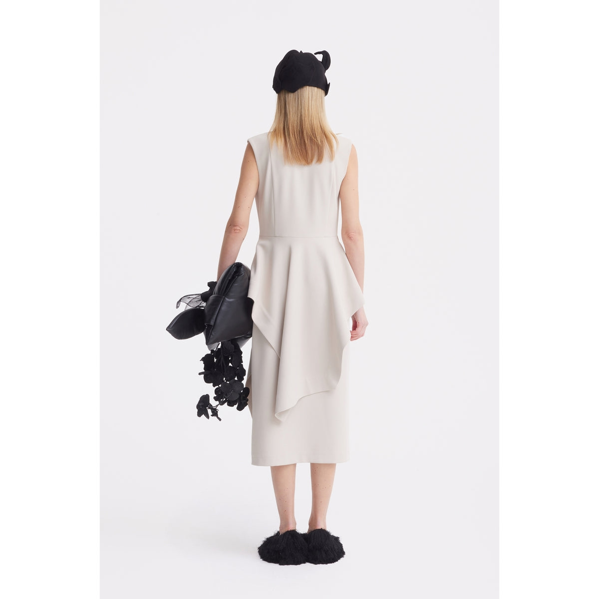 A person with straight blonde hair faces away, wearing the JULIA ALLERT Architectural Draped Midi Dress in beige, paired with a black beret, black fuzzy shoes, and holding black accessories. The background is white.