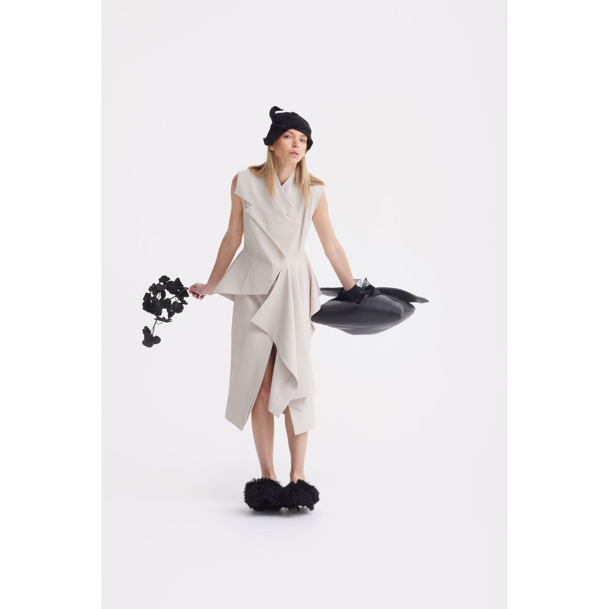 A woman wearing the JULIA ALLERT Architectural Draped Midi Dress in beige stands against a white background, accessorized with a black hat, sculptural abstract black bag, and black fluffy shoes.