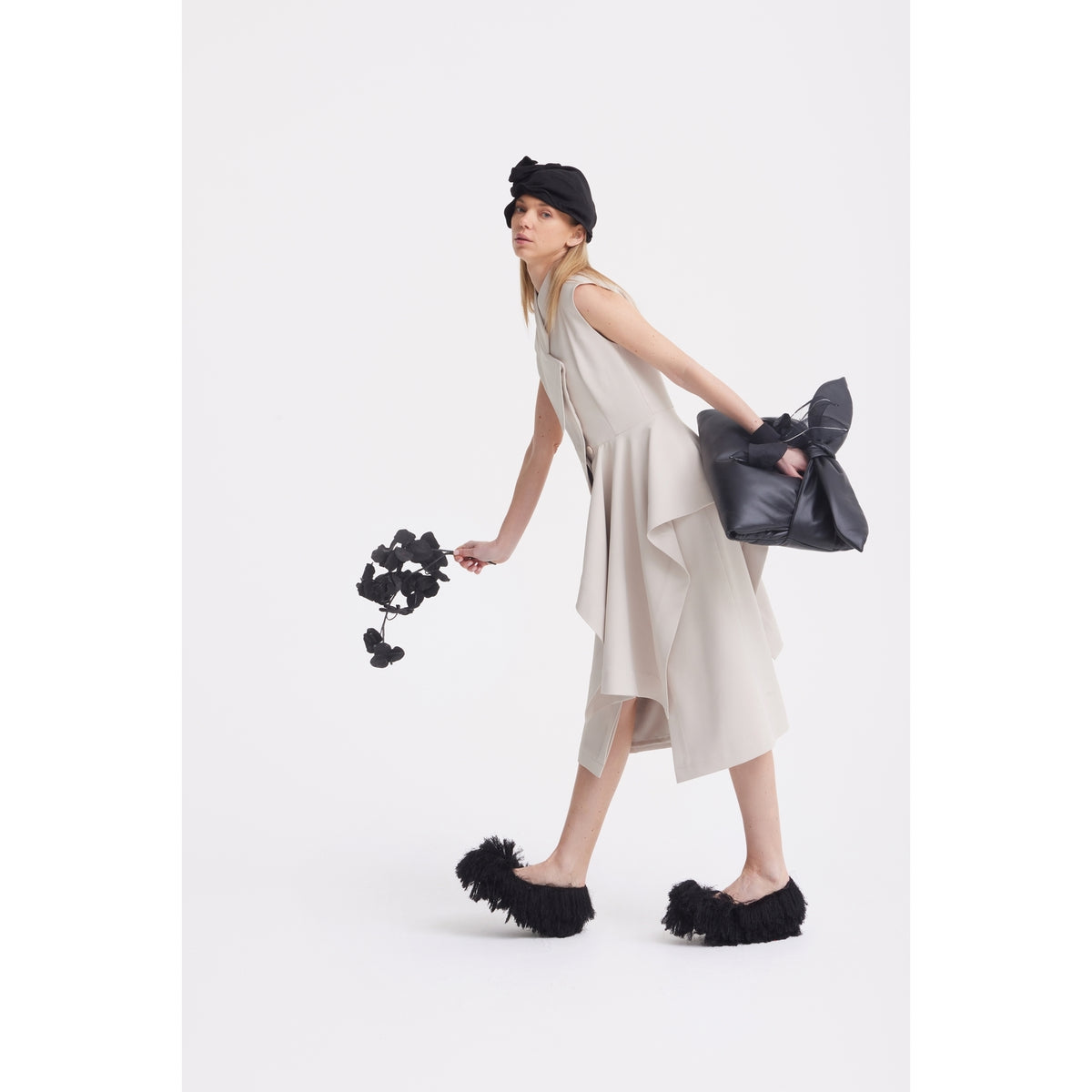 A woman wears the JULIA ALLERT Architectural Draped Midi Dress in beige with black fluffy shoes, a black headpiece, artificial black flowers, and a large black bow accessory, posing against a white background.