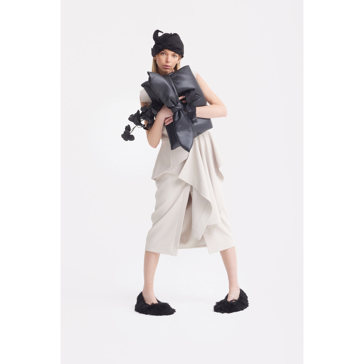 A woman poses in the JULIA ALLERT Architectural Draped Midi Dress in beige, styled with a large black bow, black slippers, and a black hat. A plain white background highlights her sculptural, avant-garde look.