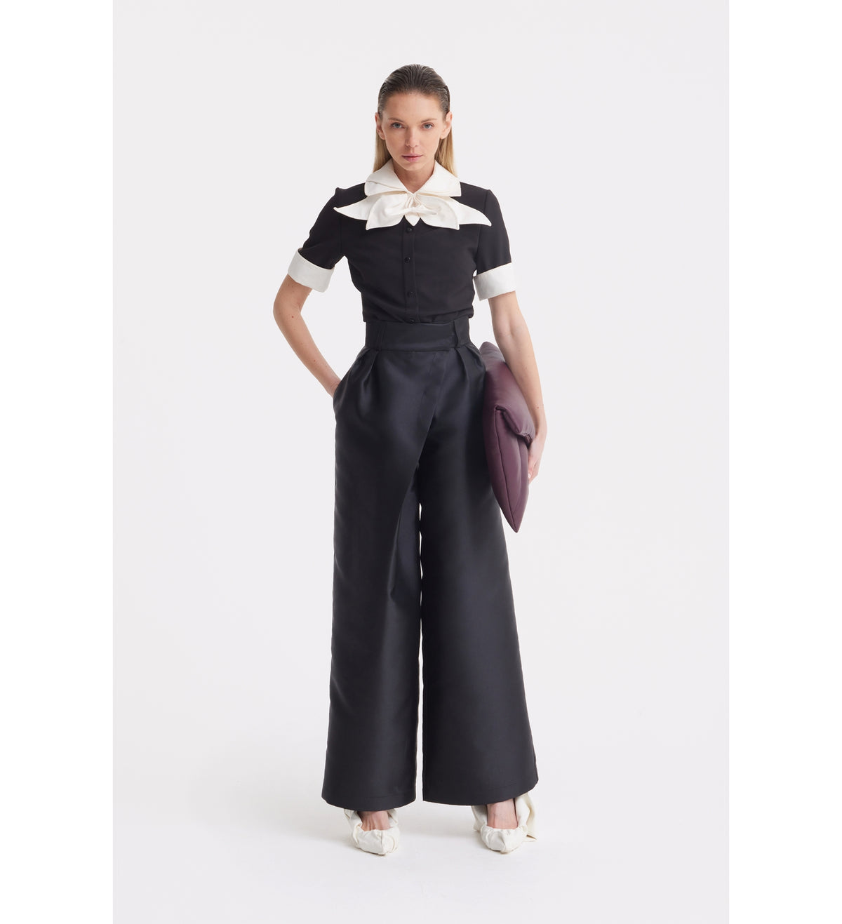 A woman stands against a white background wearing the JULIA ALLERT Short Sleeve Formal Bow Blouse in black, featuring a large white contrast bow collar and cuffs, paired with high-waisted wide-leg black pants, white shoes, and holding a purple clutch.