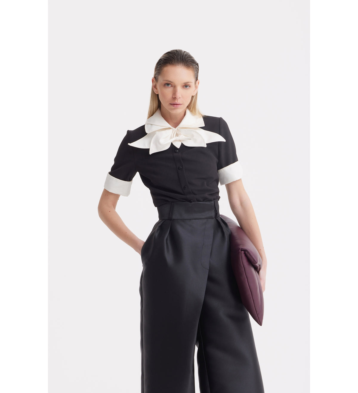A woman with straight blonde hair wears the JULIA ALLERT Short Sleeve Formal Bow Blouse in black, featuring a contrast bow collar and white cuffs, paired with high-waisted black pants. She holds a dark purple clutch against a plain white background.