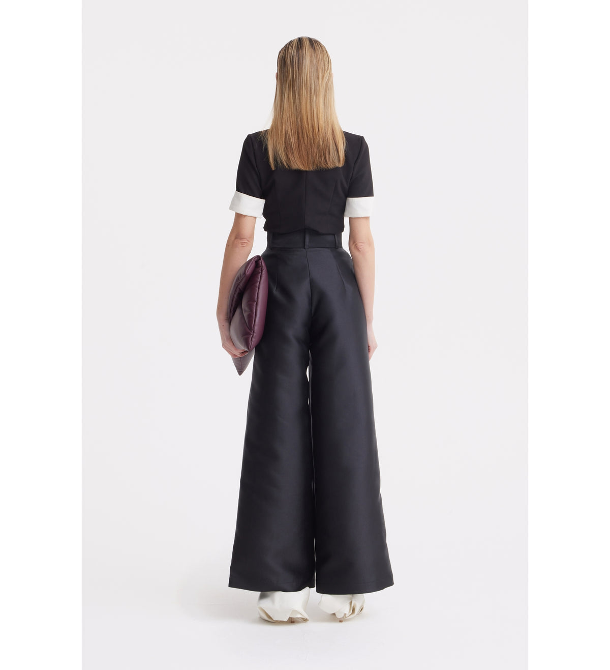 A person with straight, shoulder-length hair stands facing away, wearing a JULIA ALLERT Short Sleeve Formal Bow Blouse - Black with a contrast bow collar, high-waisted wide-leg black pants, white shoes, and holding a large dark clutch.