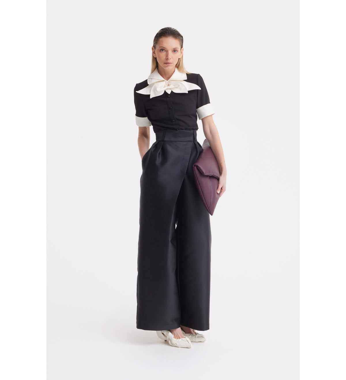 A woman poses against a white background wearing a JULIA ALLERT Short Sleeve Formal Bow Blouse in black, paired with high-waisted wide-leg black pants, white shoes, and holding a large purple clutch.