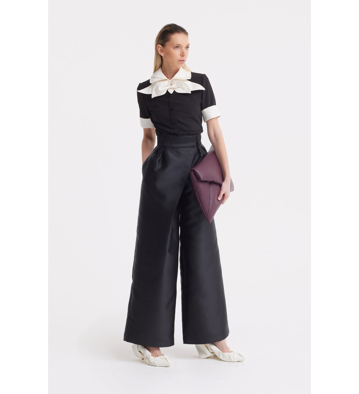 A woman stands against a white background wearing a JULIA ALLERT Short Sleeve Formal Bow Blouse in black with a large contrast bow collar, high-waisted wide-leg black pants, white shoes, and holding a large purple clutch.