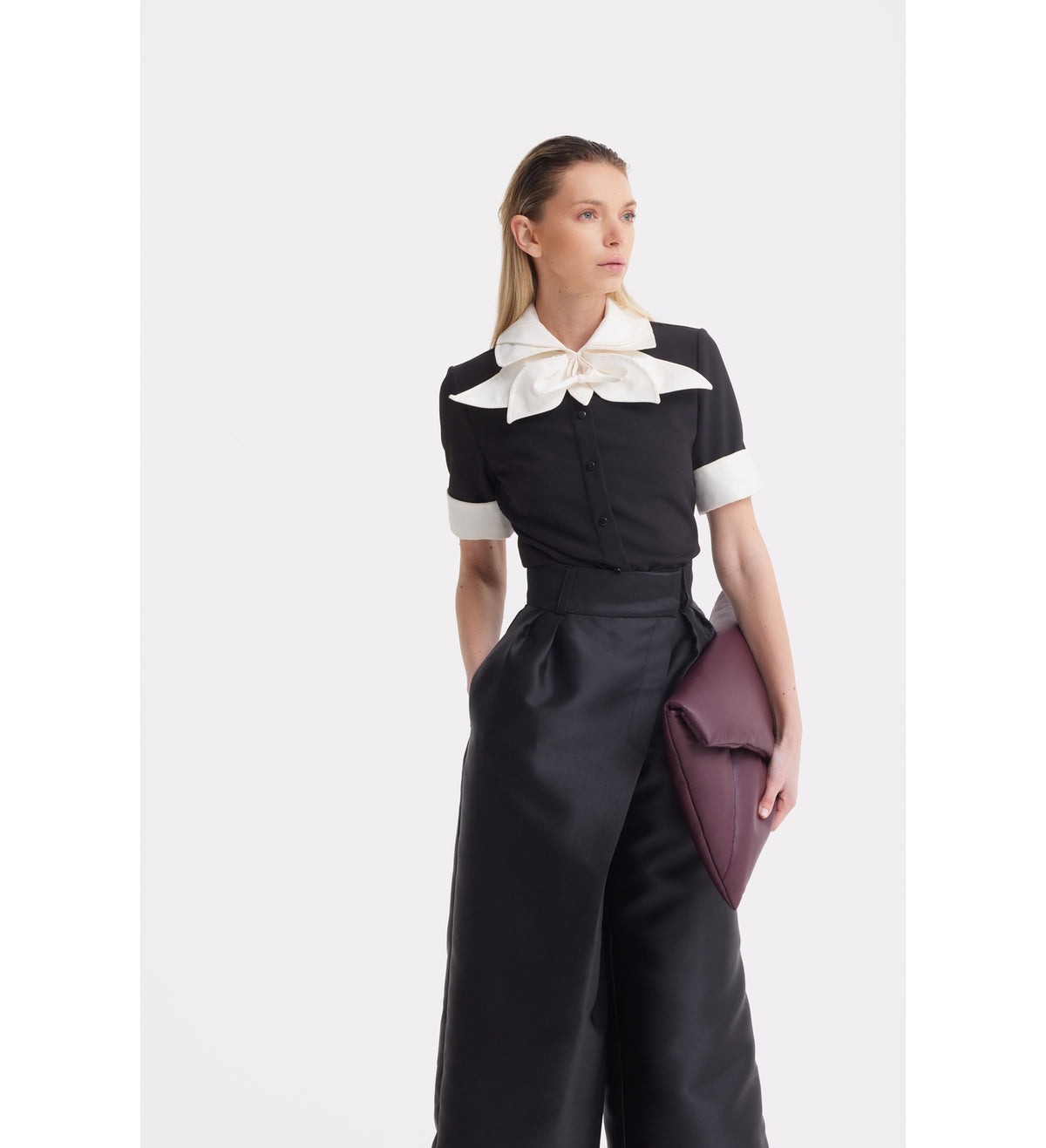 A woman stands against a plain background in a JULIA ALLERT Short Sleeve Formal Bow Blouse - Black, styled with wide-leg black pants and holding a large burgundy clutch under her arm.