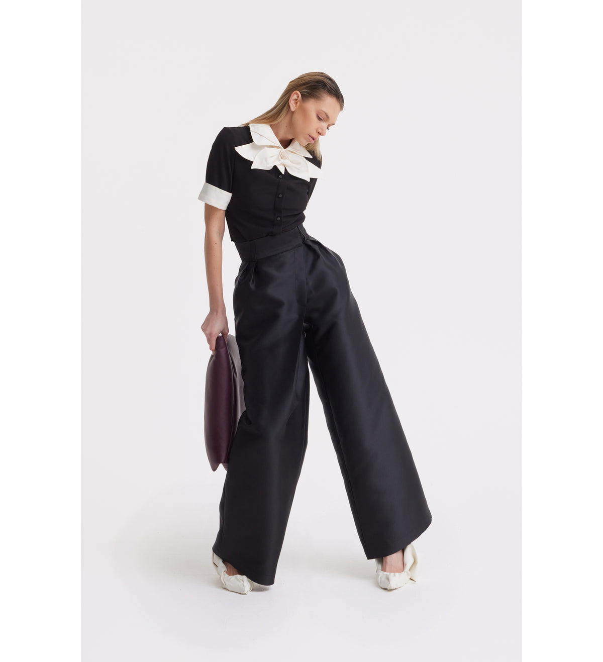 A woman poses against a white background wearing the JULIA ALLERT Short Sleeve Formal Bow Blouse - Black, styled with wide-leg black trousers, white heels, and holding a dark oval-shaped clutch.