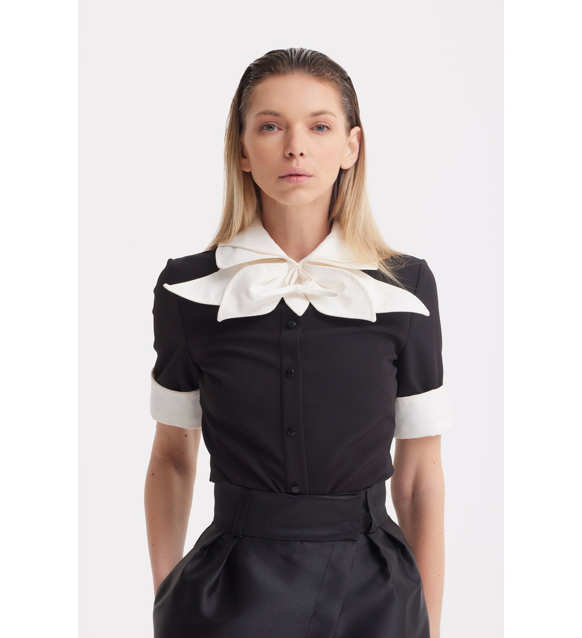 A woman with straight blonde hair models the JULIA ALLERT Short Sleeve Formal Bow Blouse - Black, featuring a contrast bow collar and white cuffs, paired with high-waisted black trousers against a plain white background.