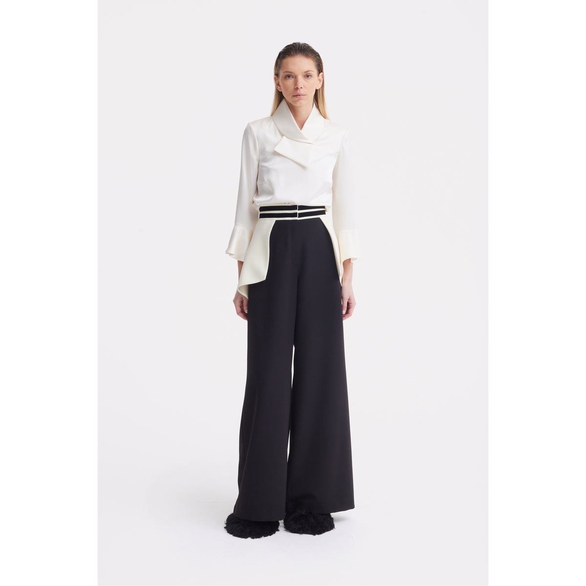 A woman with straight hair and a neutral expression wears JULIA ALLERT Statement Waist Wide-Leg Trousers in black and a white long-sleeve blouse, standing against a plain white background.
