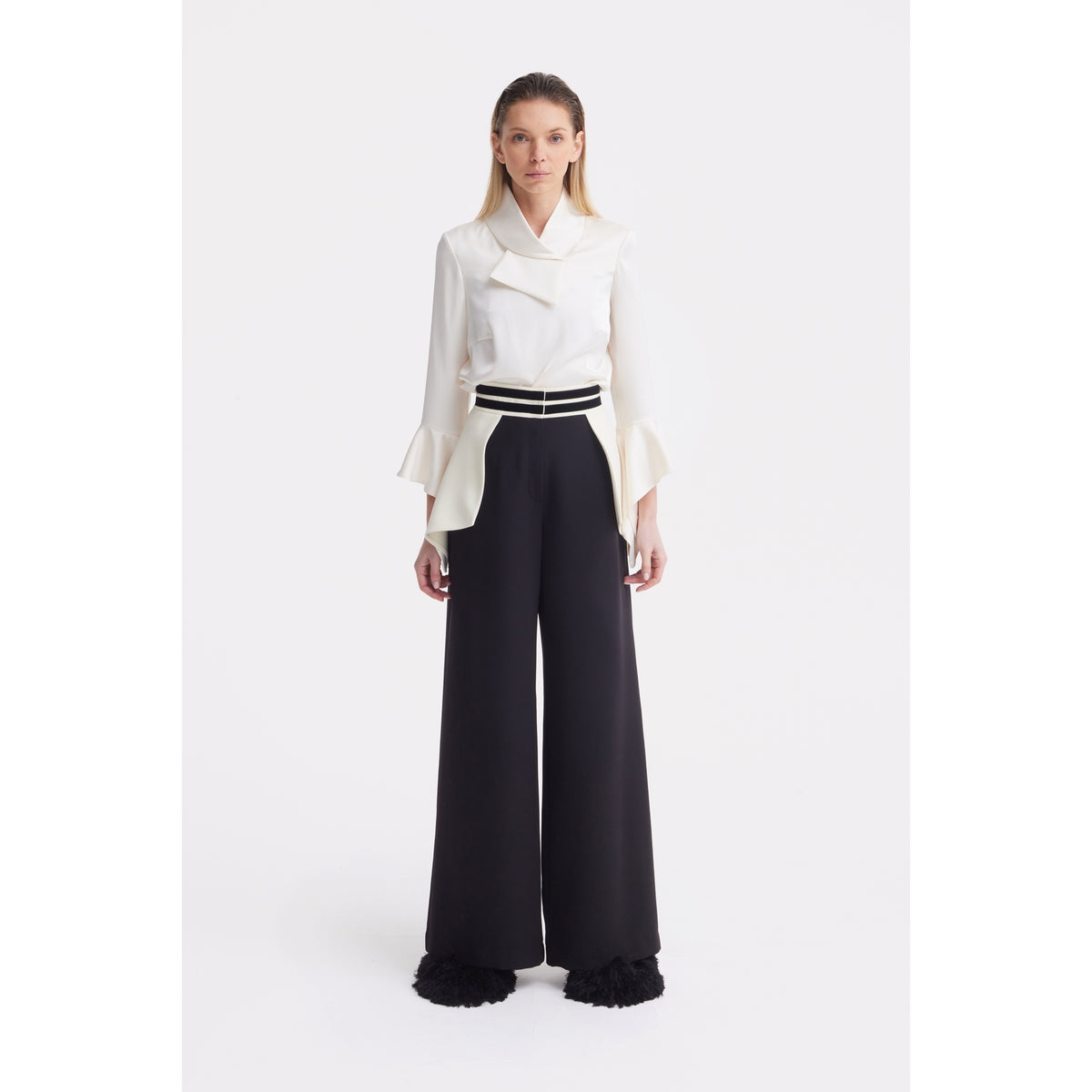 A woman stands facing forward, wearing a JULIA ALLERT Asymmetric Collar Satin Blouse in ivory and high-waisted, wide-leg black trousers with a striped belt, set against a plain white background.