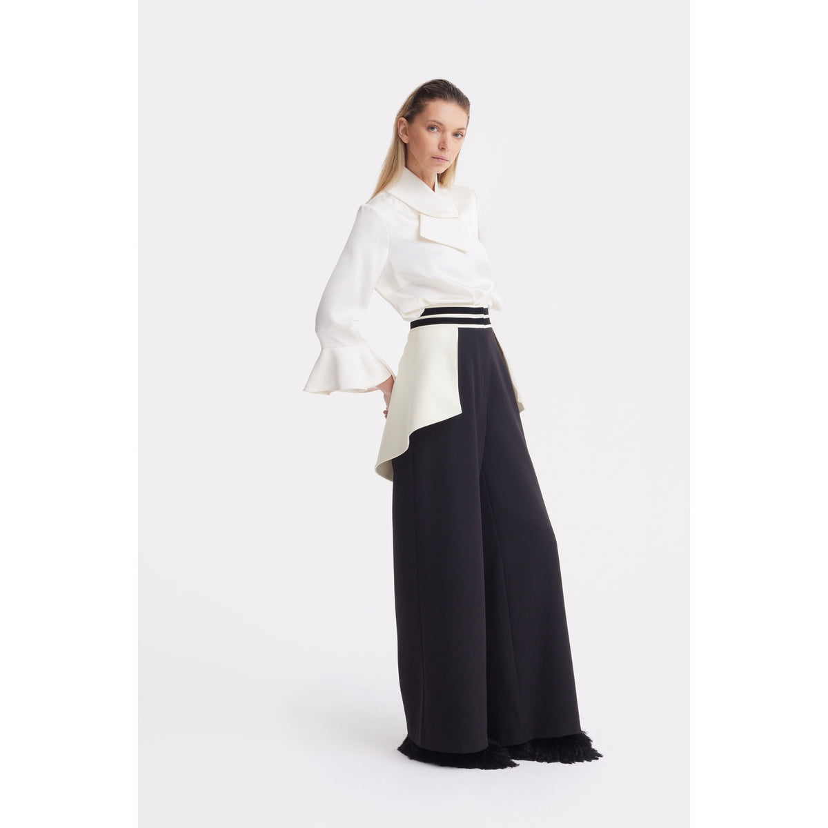 A woman poses against a white background wearing JULIA ALLERT Statement Waist Wide-Leg Trousers in black, featuring an asymmetrical white overlay at the high-waisted, tailored-fit waist.