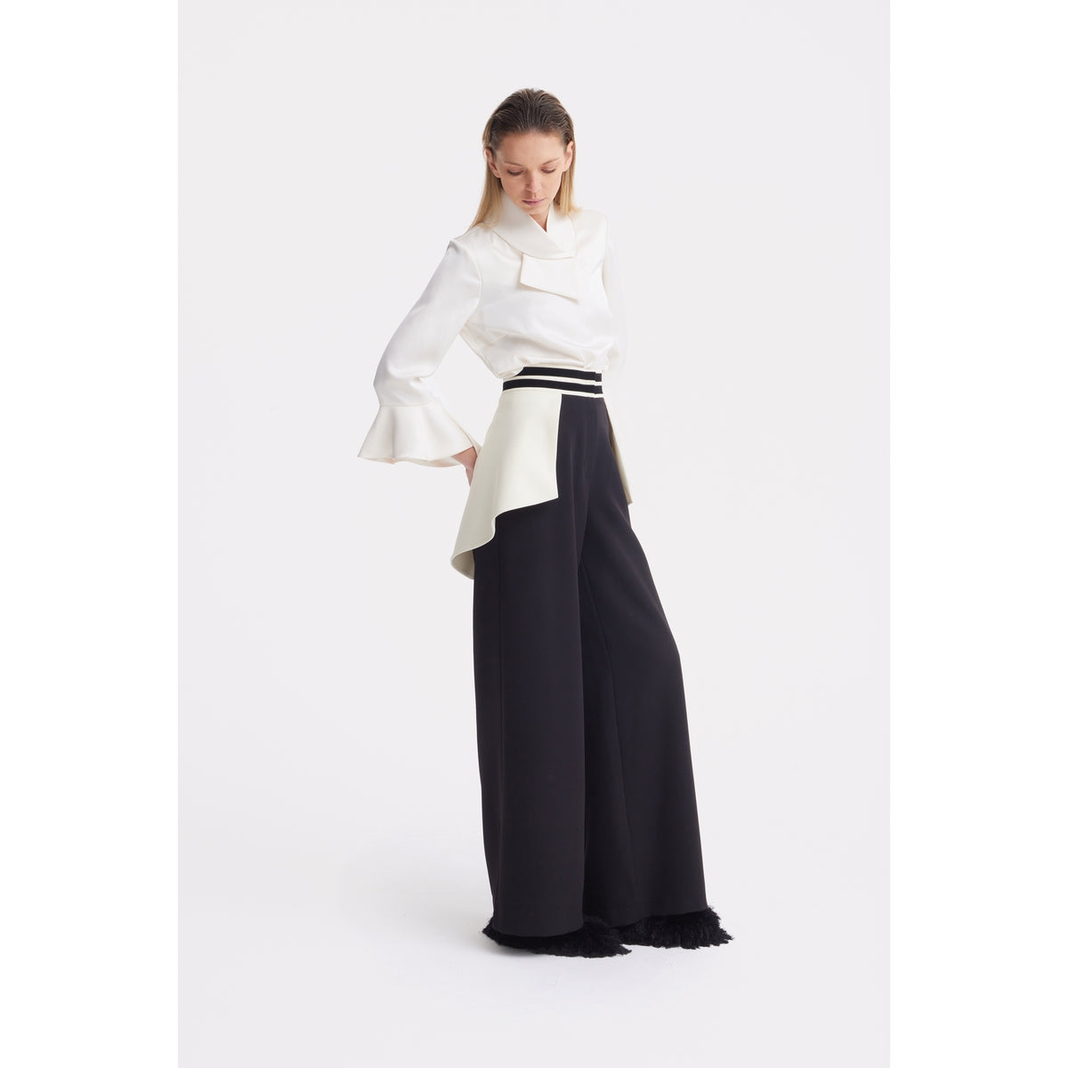 A woman wears JULIA ALLERT Statement Waist Wide-Leg Trousers in black, featuring a white asymmetrical overskirt and fringe hem. She pairs them with a white blouse with flared sleeves and a large collar, looking down with hands at her sides.