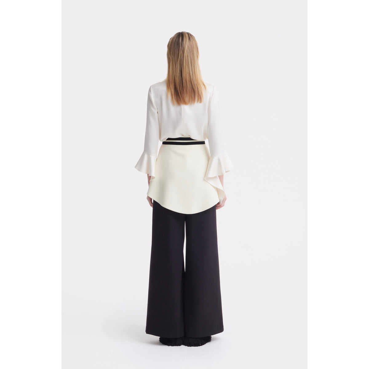 A person stands facing away, wearing a JULIA ALLERT Asymmetric Collar Satin Blouse in ivory with flared sleeves, styled over black wide-leg pants, set against a plain white background.