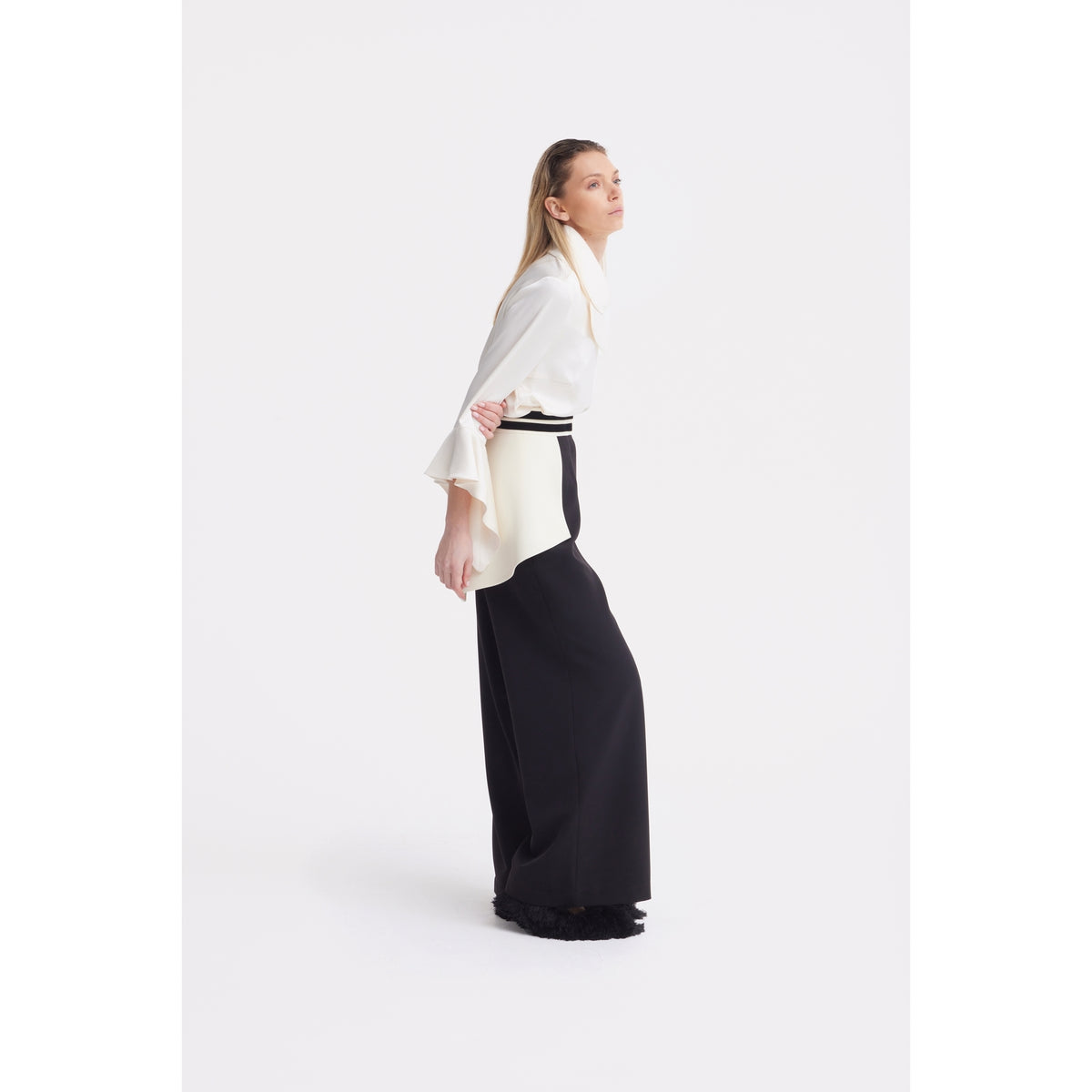 A woman stands in profile against a white background, wearing the JULIA ALLERT Asymmetric Collar Satin Blouse in ivory with flared sleeves and high-waisted, wide-leg black pants. Her hair is straight and she has a calm, neutral expression.