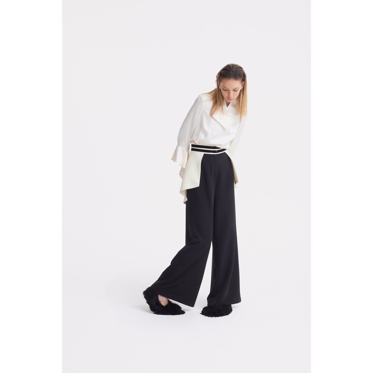A woman stands against a white background wearing a white long-sleeve blouse, JULIA ALLERT Statement Waist Wide-Leg Trousers in black with a striped belt, and black fluffy slippers, looking down with her hands by her sides.